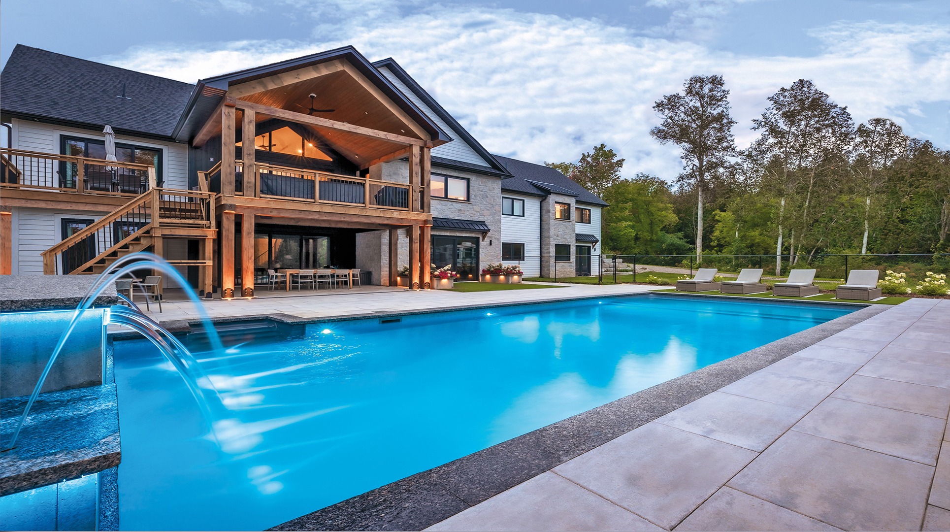 A modern house with a spacious pool and fountains, surrounded by trees and lounge chairs on a sunny day.