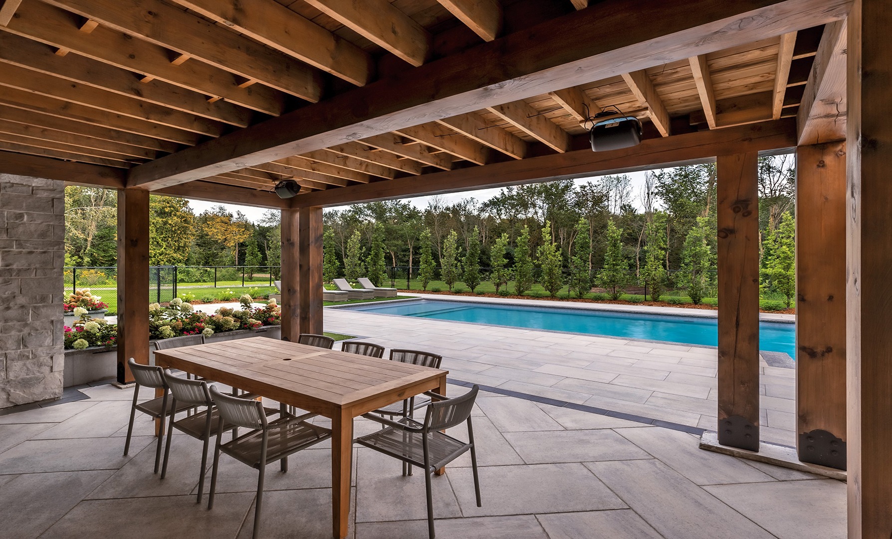 A covered patio with a wooden dining set overlooks a landscaped backyard, featuring a swimming pool surrounded by trees and neatly arranged flowers.