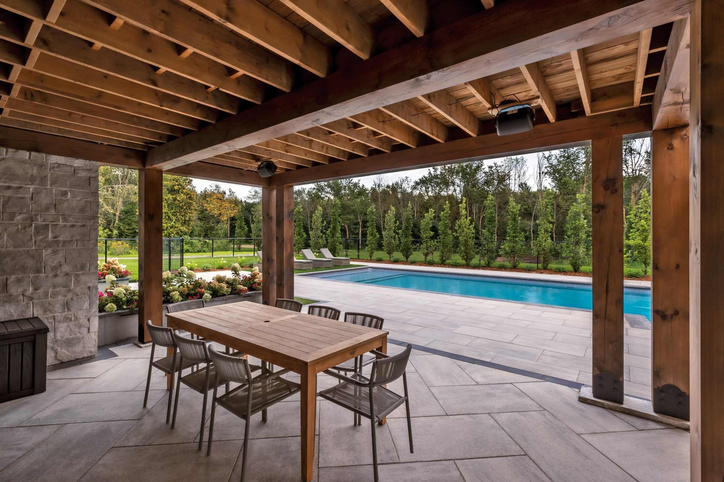 A covered patio with wooden beams overlooks a swimming pool. Outdoor dining table and chairs. Lush greenery surrounds the area, creating a serene atmosphere.