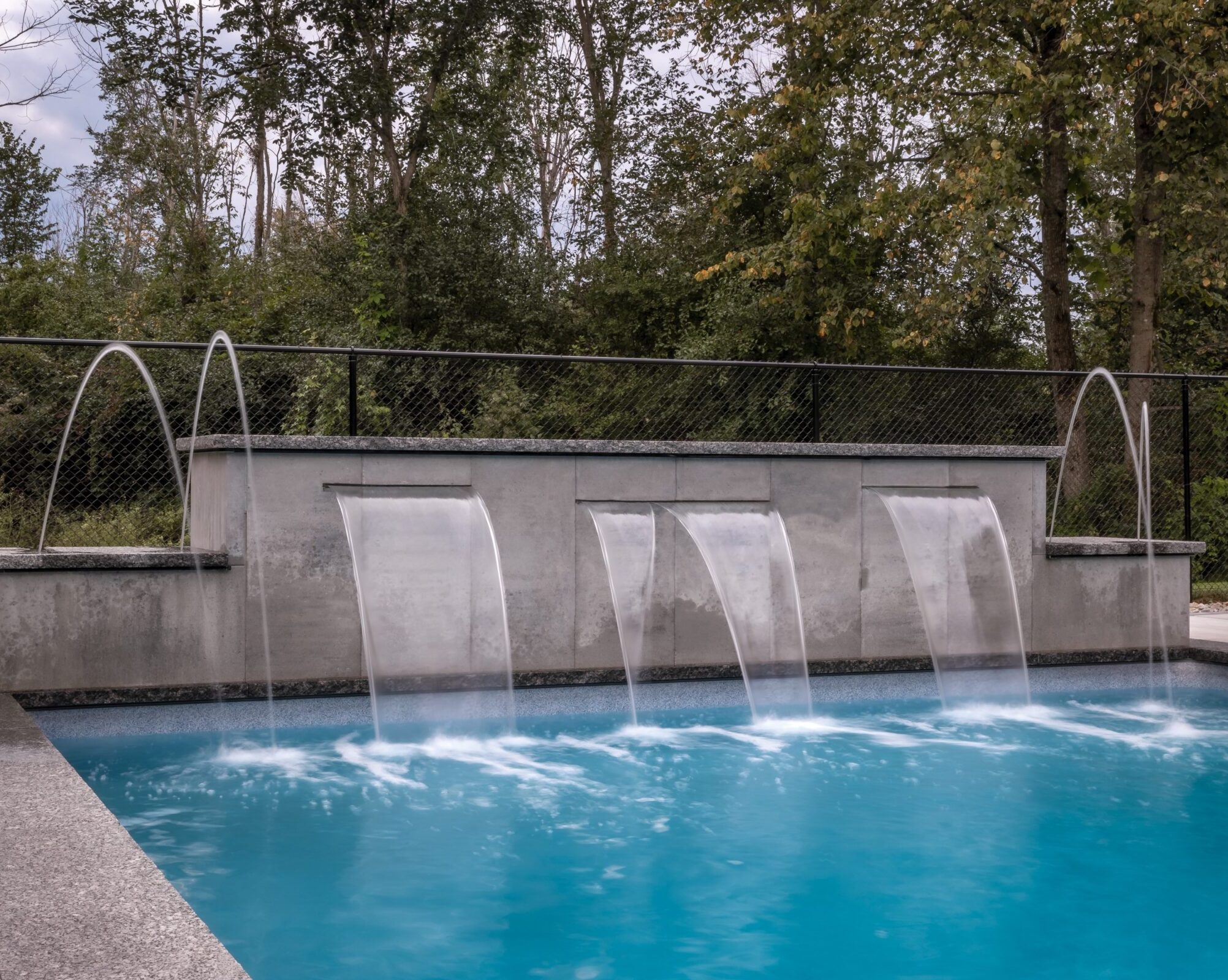 A modern swimming pool with cascading water features and a sleek design, set against a backdrop of lush green trees on a cloudy day.