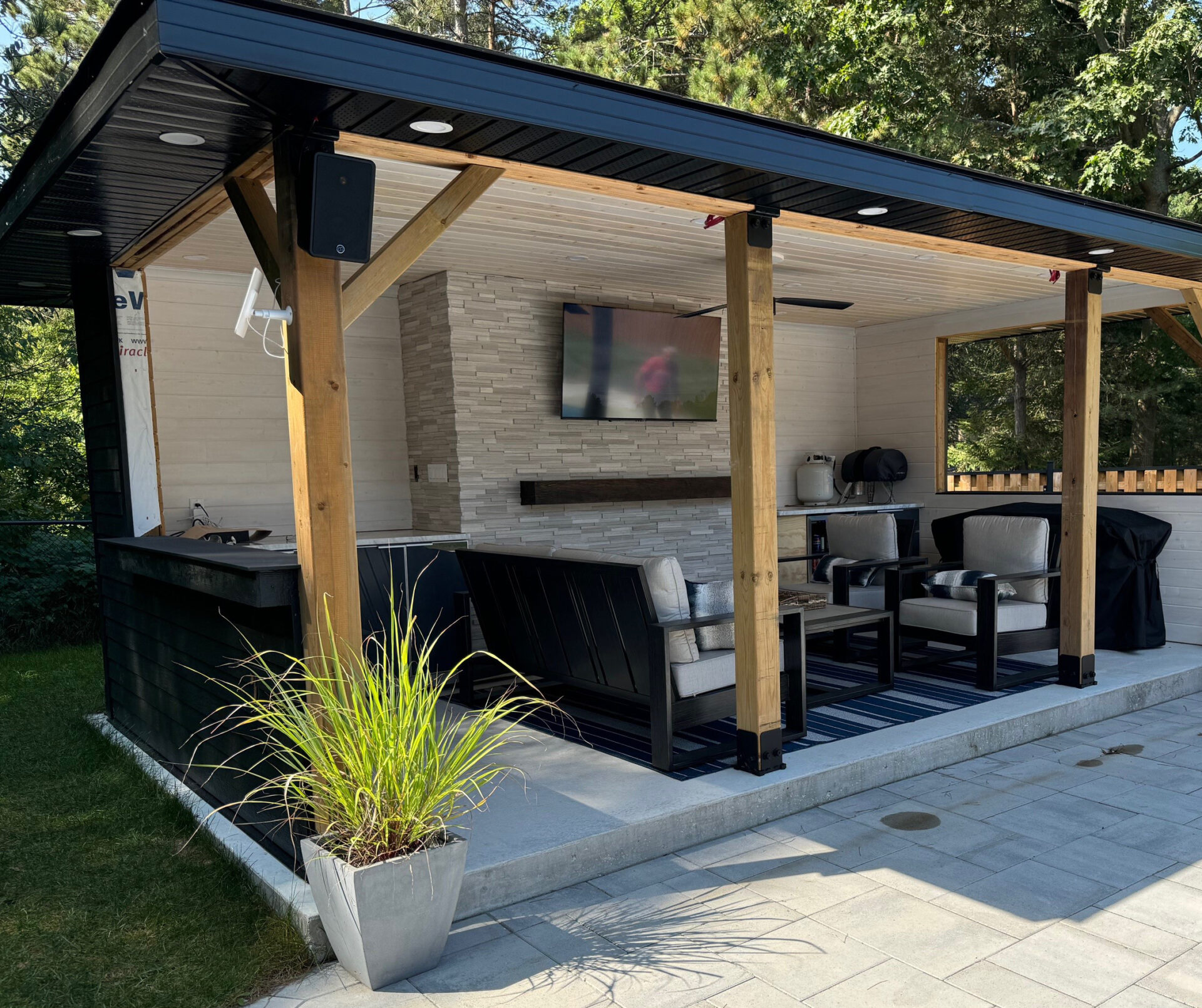 A cozy outdoor patio has modern seating, TV, barbecue grill, and potted plant, surrounded by lush greenery and wooden beams for support.