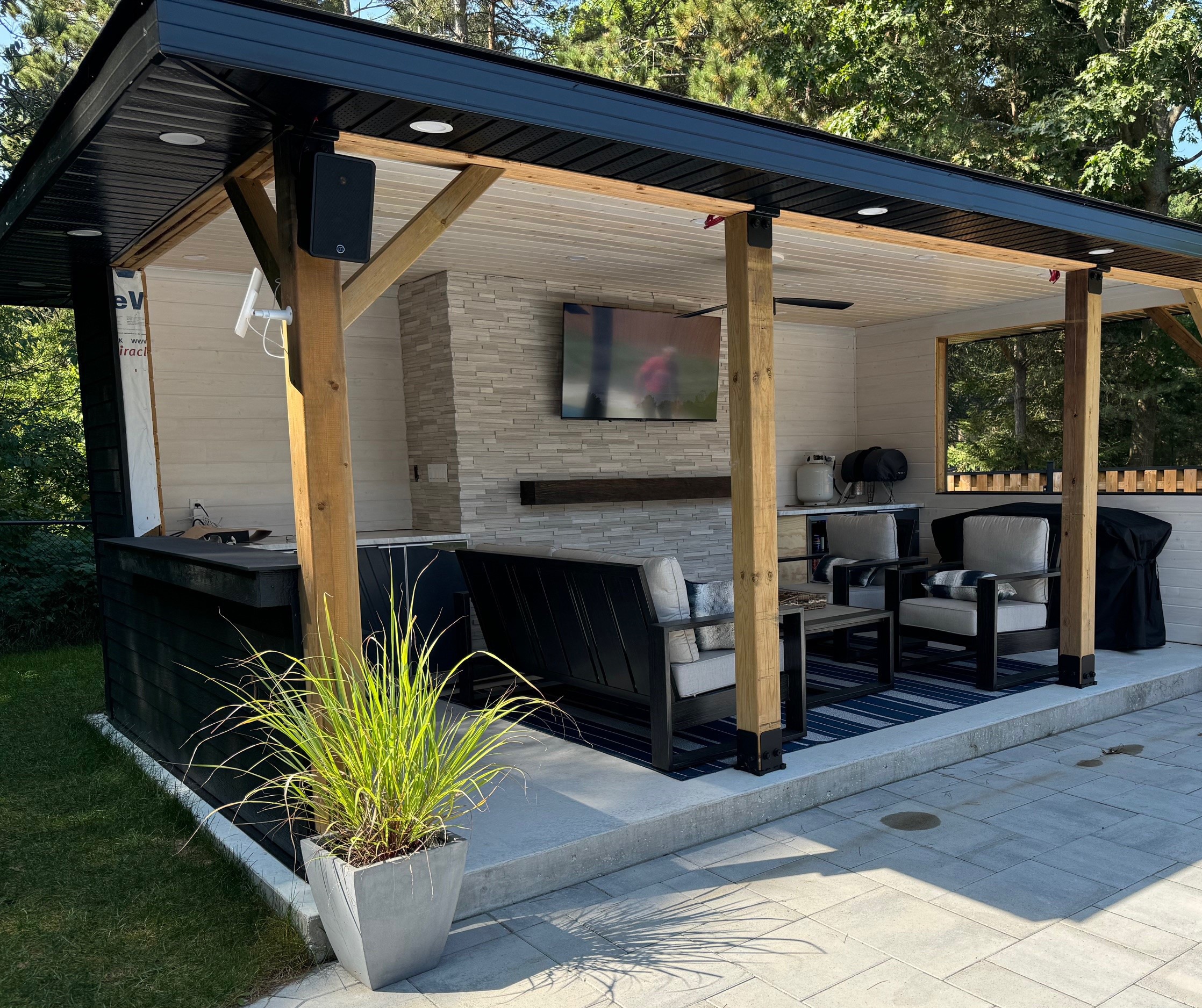 A cozy outdoor patio has modern seating, TV, barbecue grill, and potted plant, surrounded by lush greenery and wooden beams for support.