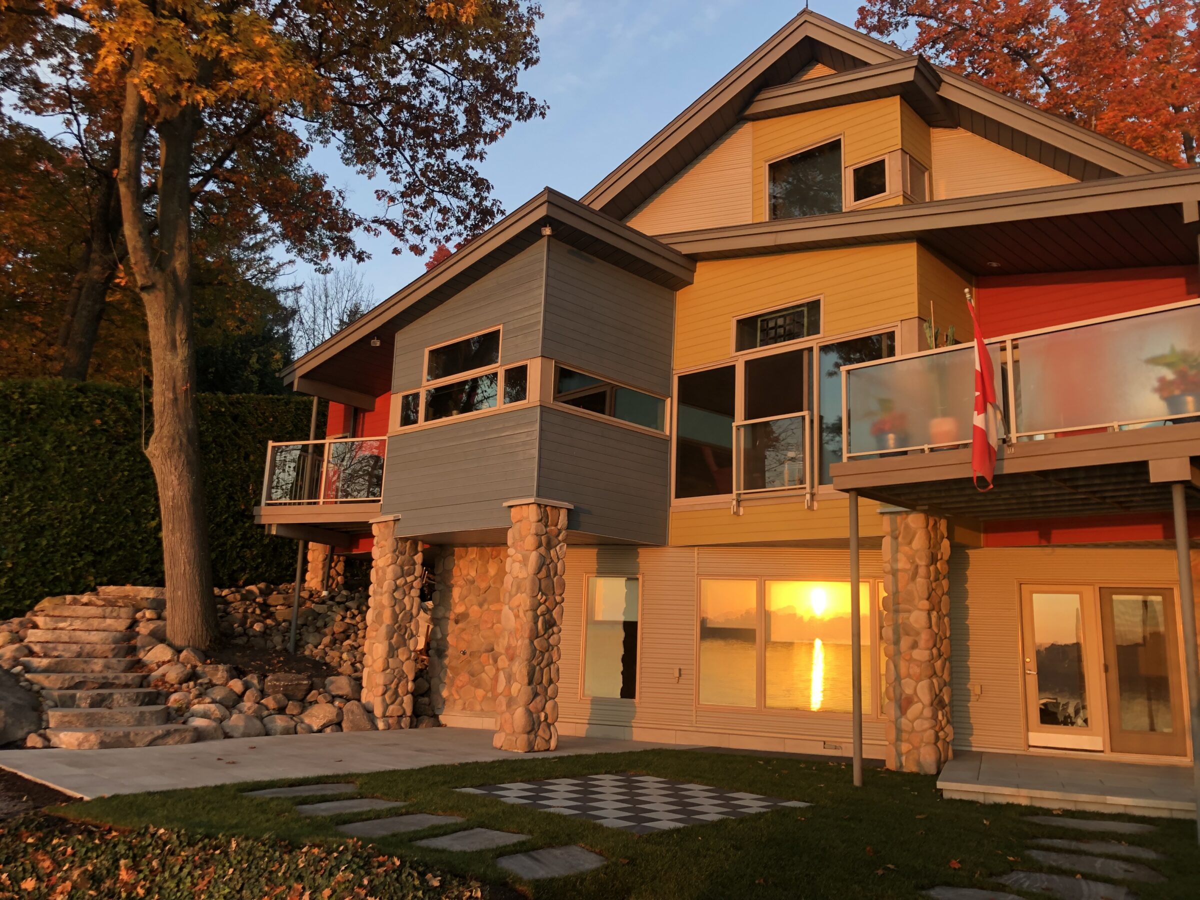Modern house with large windows and stone pillars, set among autumn trees. Sunlight reflects brightly, highlighting sleek design and peaceful surroundings.