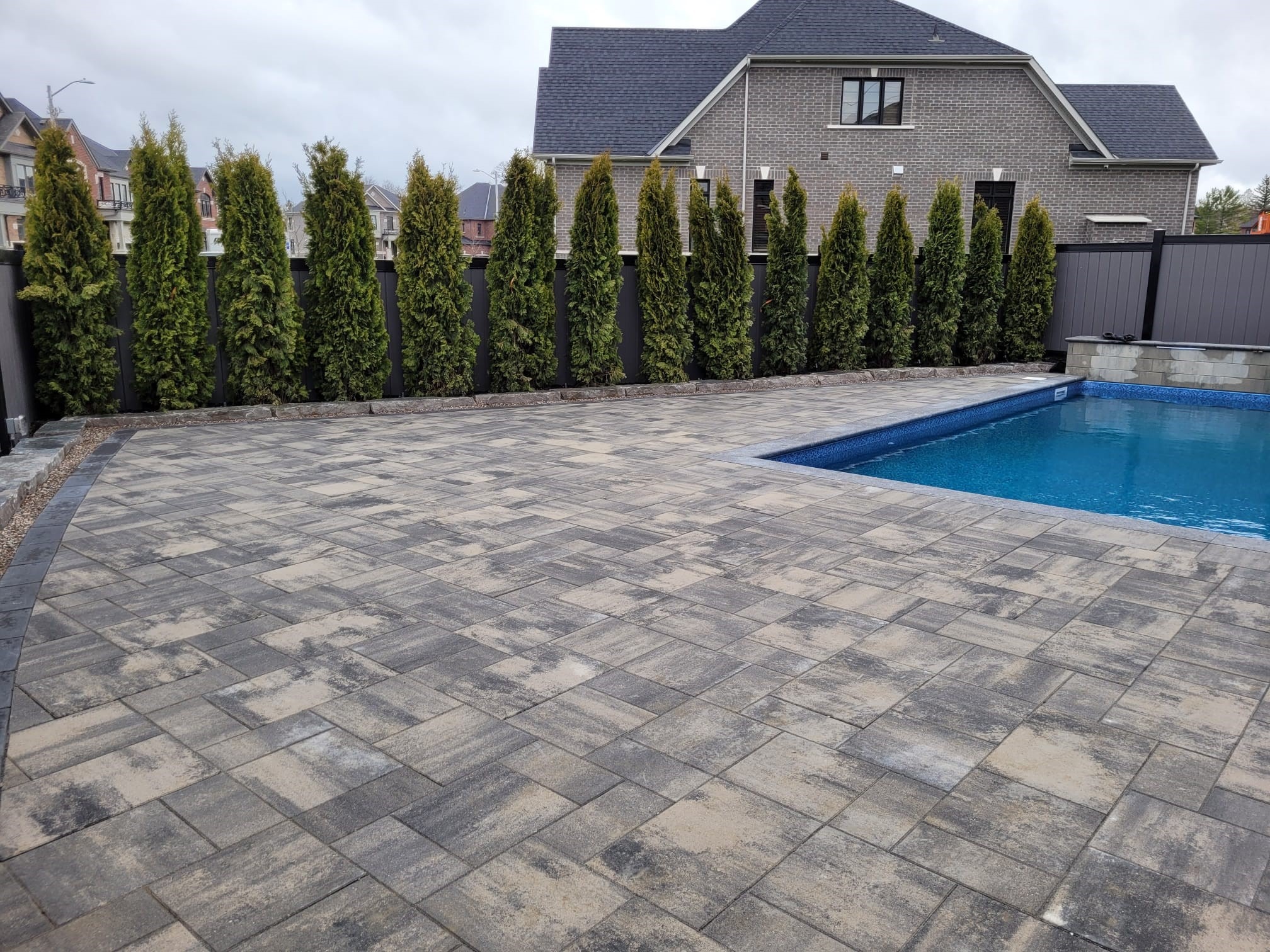 A modern house with a paved patio and pool is surrounded by tall cedar trees in a suburban neighborhood setting.