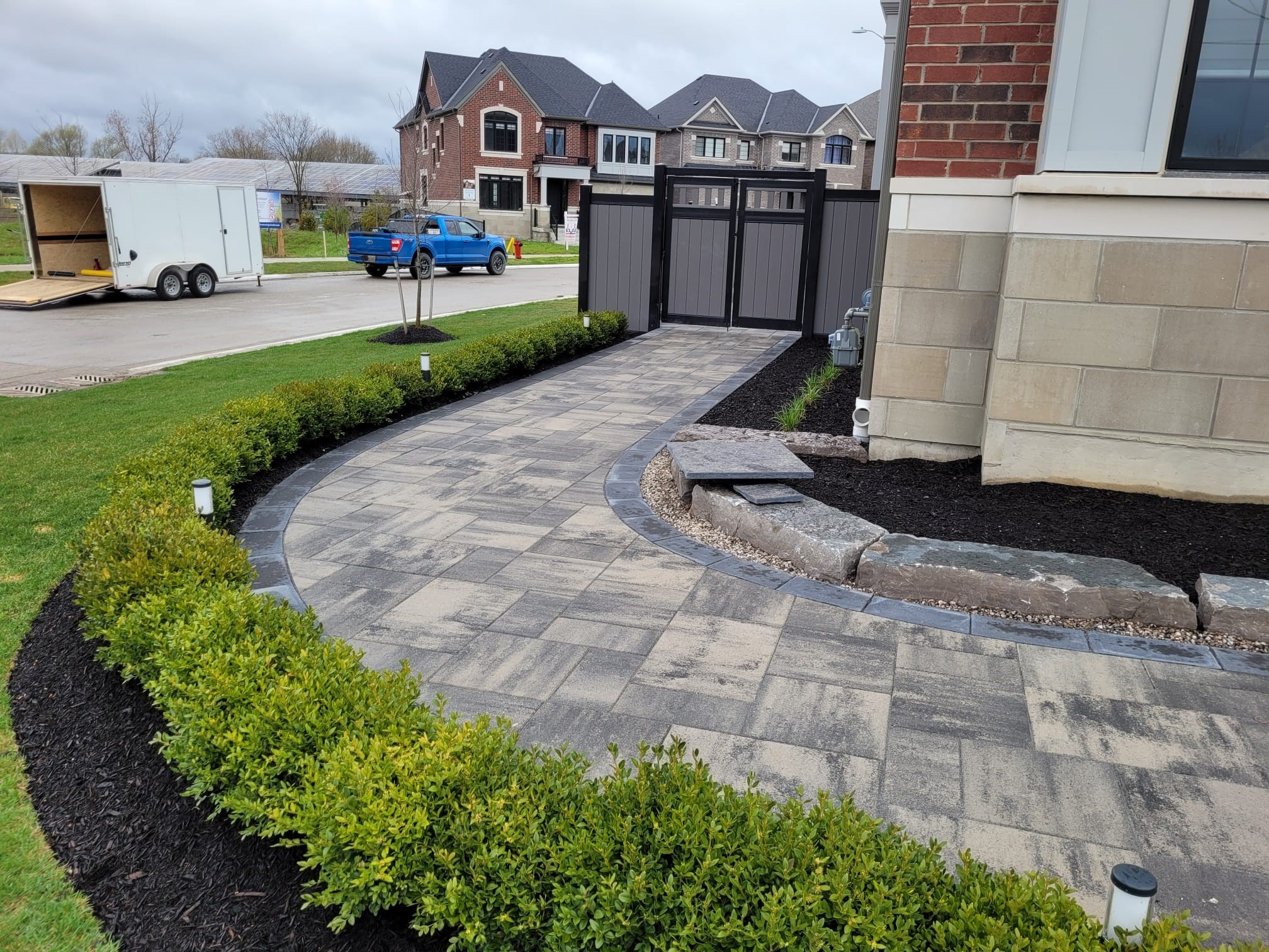 Curved stone pathway lined with green shrubs, leading to a modern suburban home. Nearby, construction trailer and blue truck on the street.