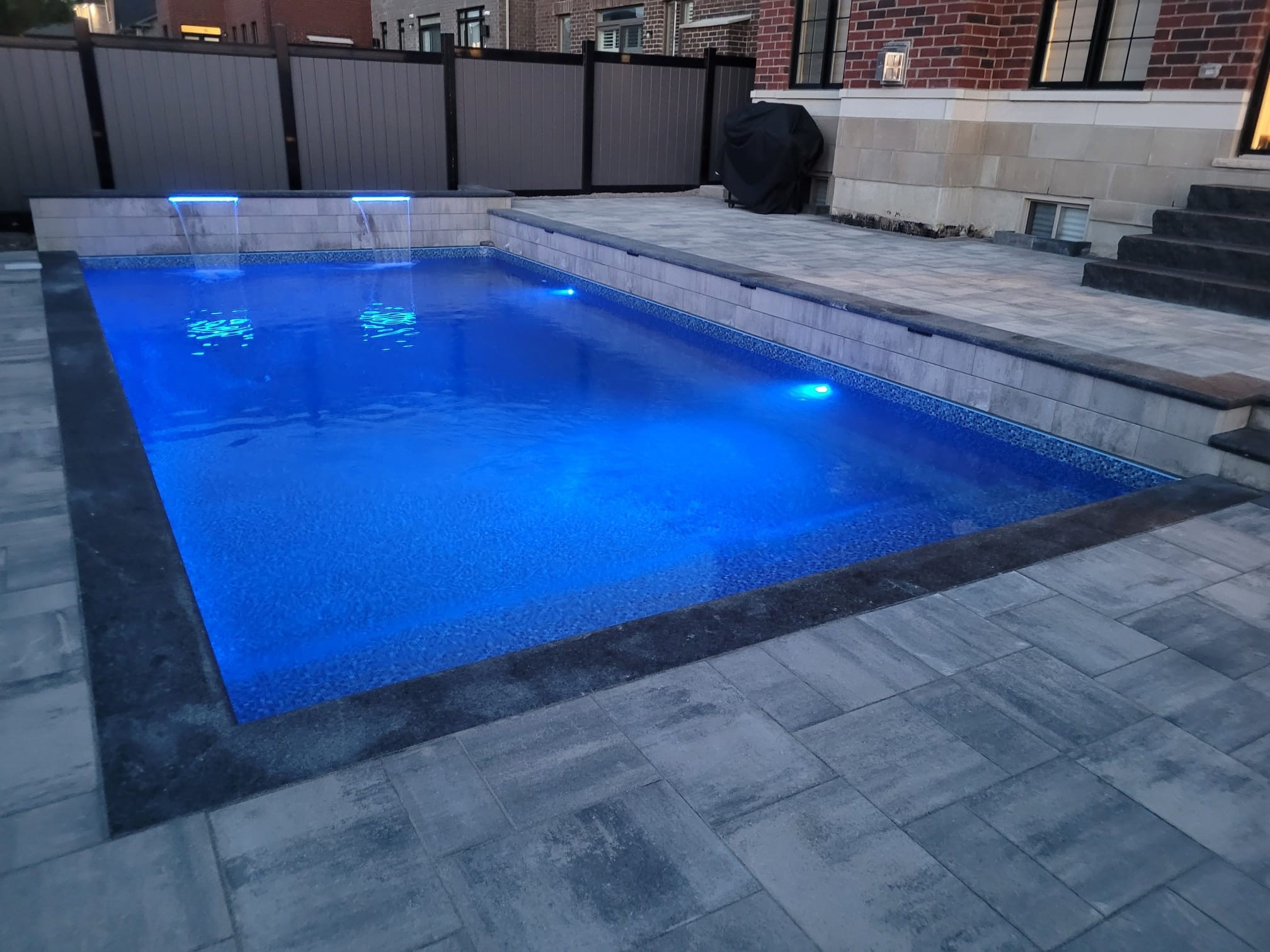 A backyard features a lit, modern pool with surrounding patio stones; adjacent brick building and fence offer privacy. No recognizable landmarks visible.