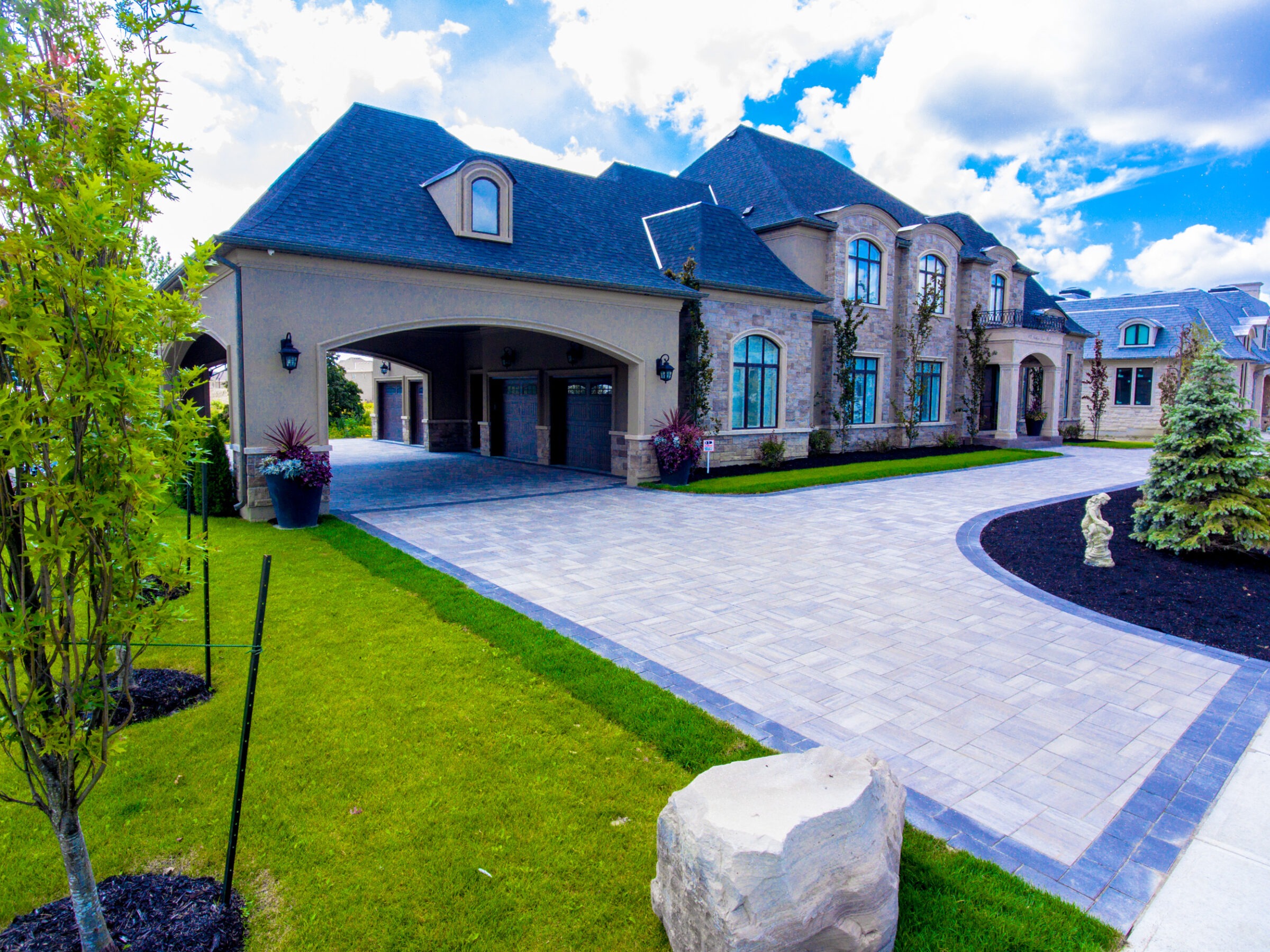 Luxurious, large stone house with manicured lawn, driveway, and ornamental trees under a vibrant blue sky with scattered clouds. No landmarks or historical buildings visible.