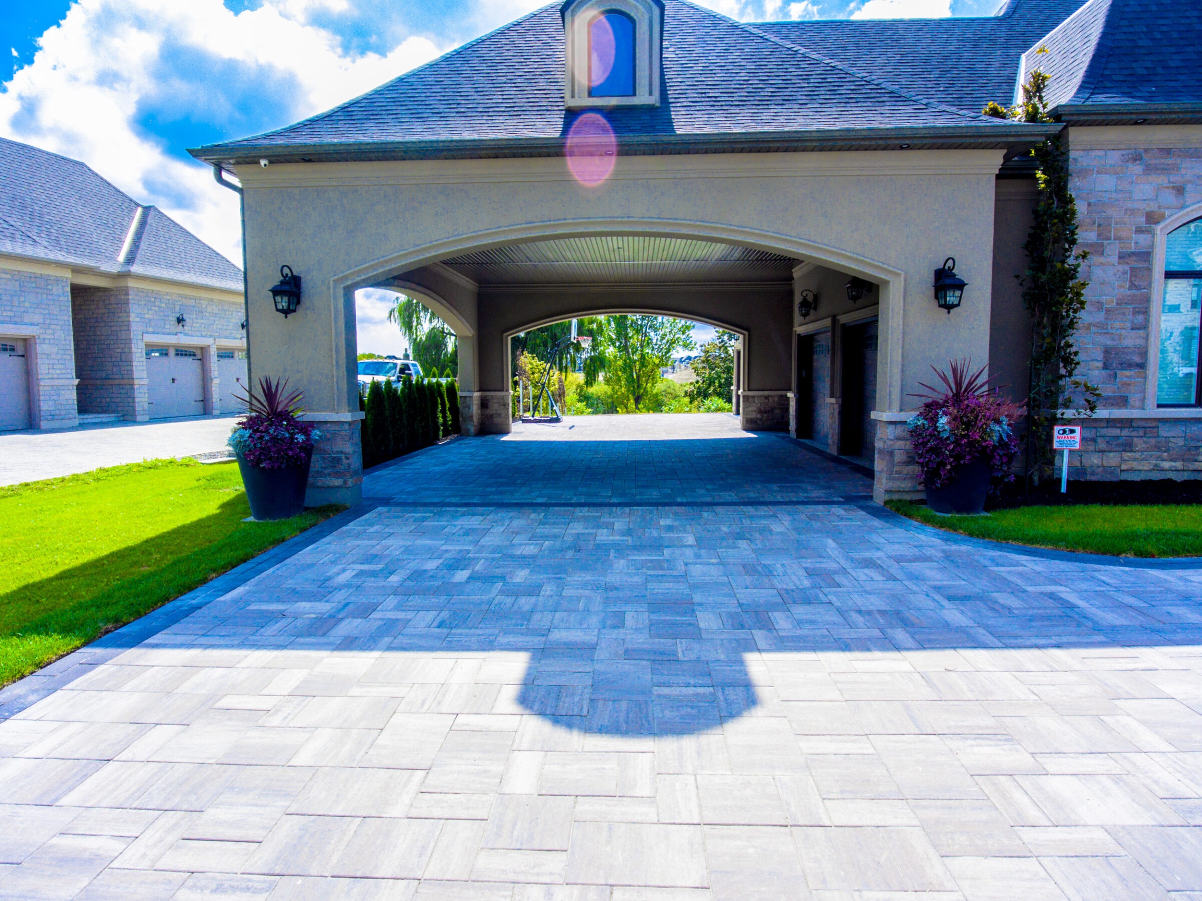 Spacious driveway leads to a modern house with an arched entryway. Lush greenery and potted plants accentuate the neat, upscale property.