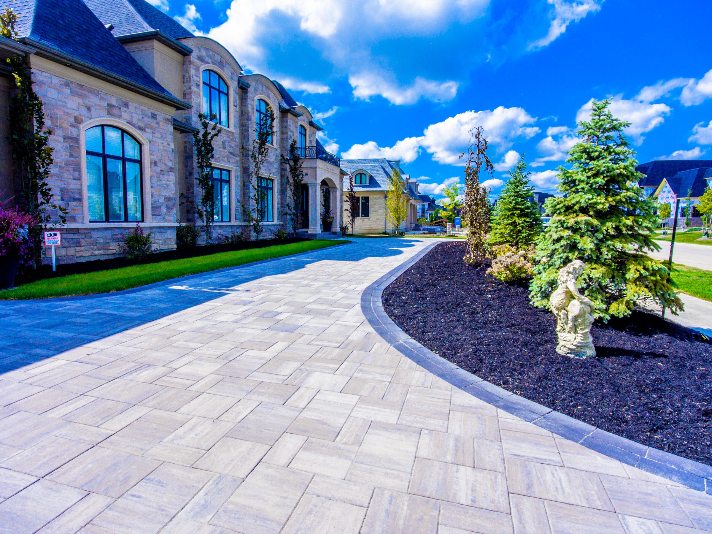 Luxurious stone mansion with arched windows, surrounded by trimmed shrubs and a paved driveway, under a bright blue sky with fluffy clouds.
