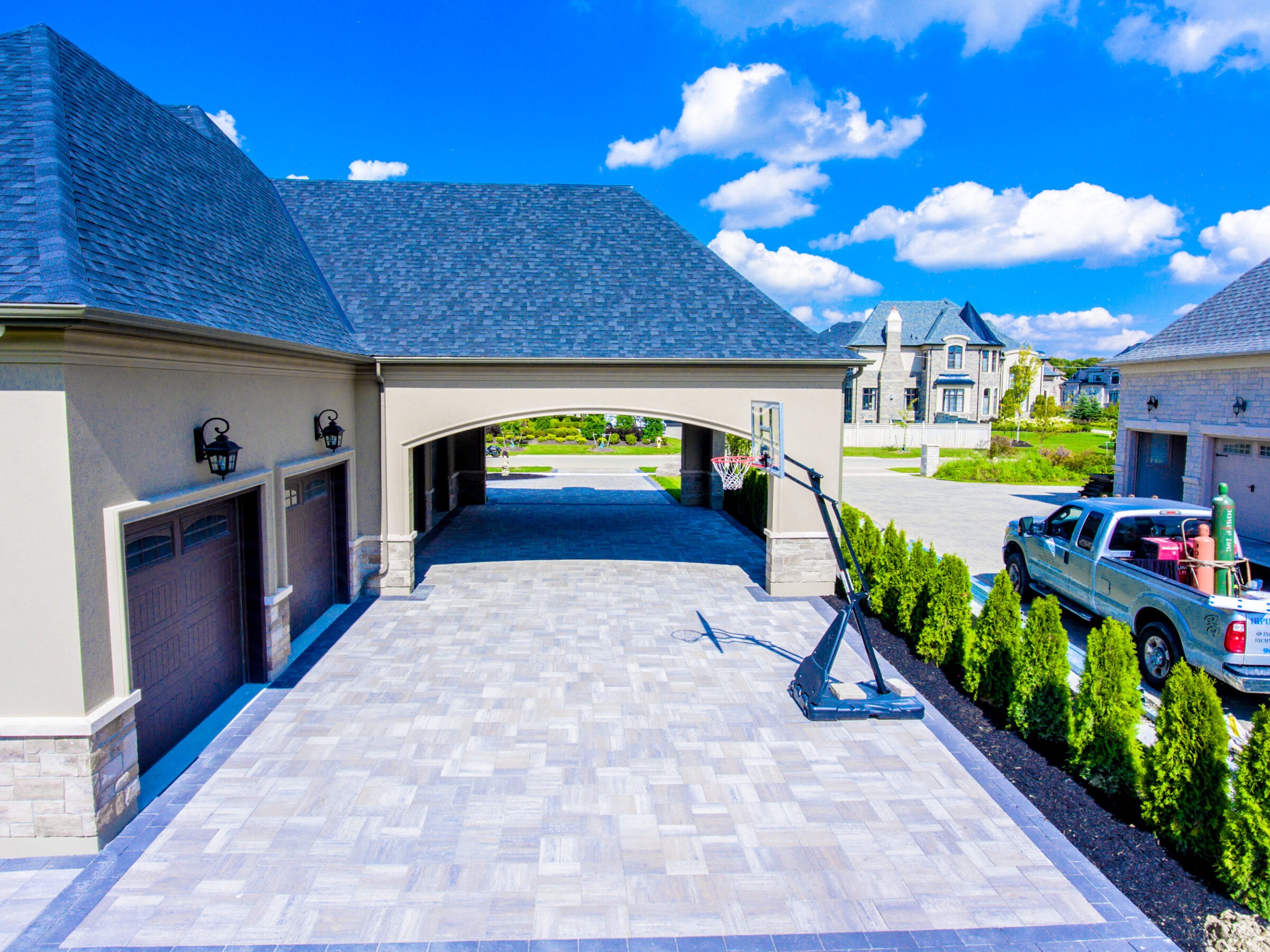 Spacious driveway with basketball hoop, surrounded by modern architecture. A truck is parked nearby, carrying items. Blue sky and clouds overhead.
