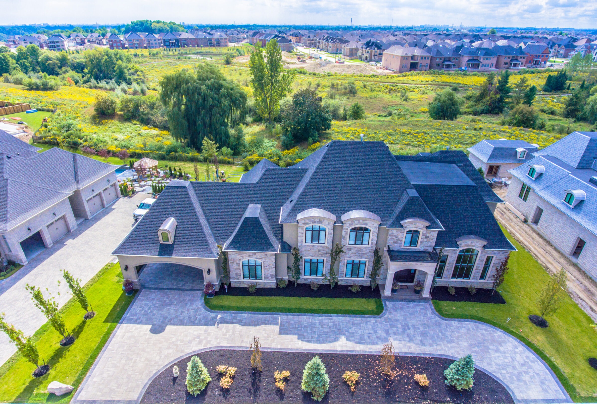 Aerial view of suburban neighborhood featuring a large, elegant house with a landscaped yard, surrounded by greenery and neighboring homes.