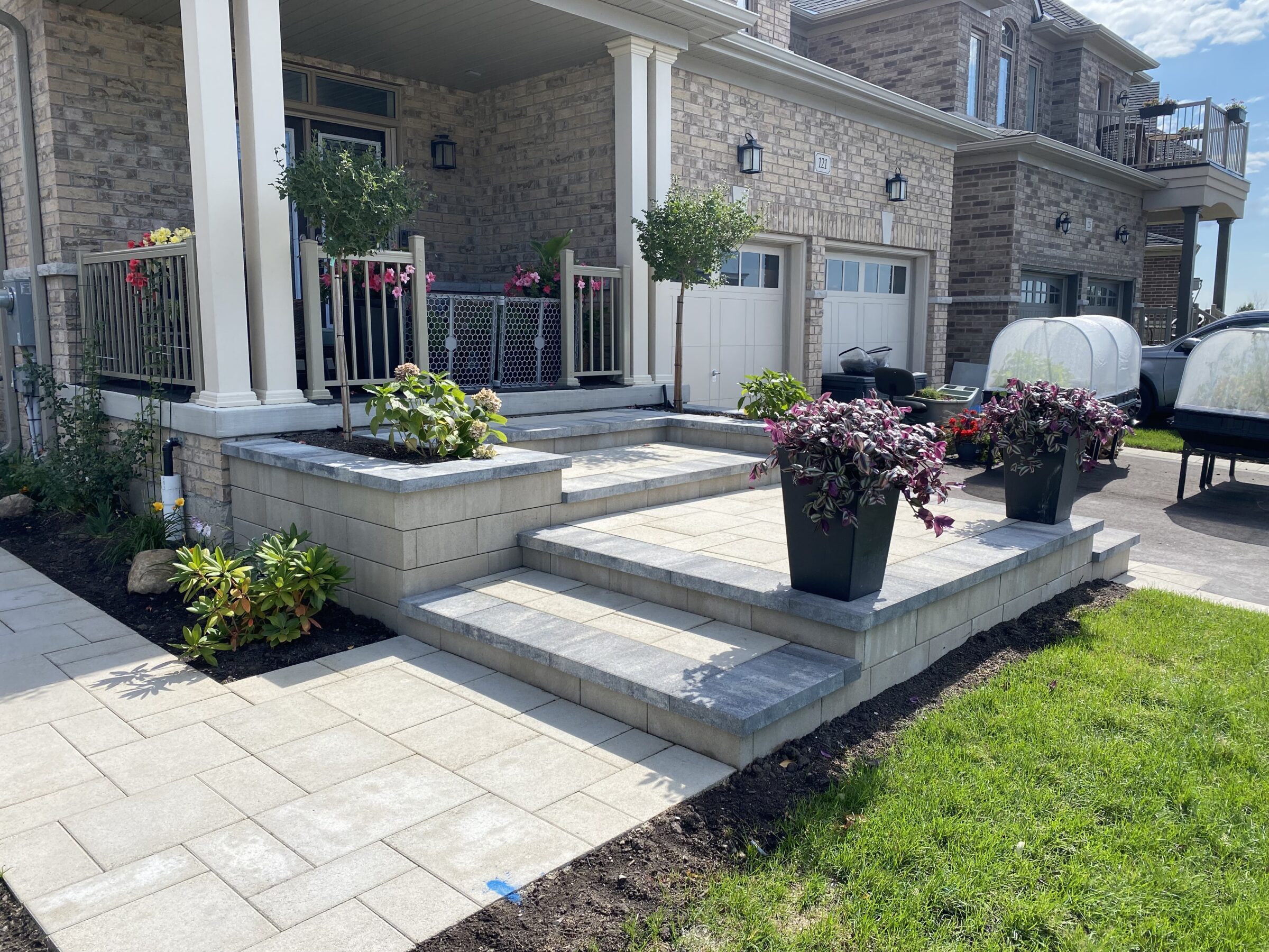 Modern house with a brick façade, landscaped front yard, and neatly arranged flower pots on a tiered porch. Driveway features cars and trailers.