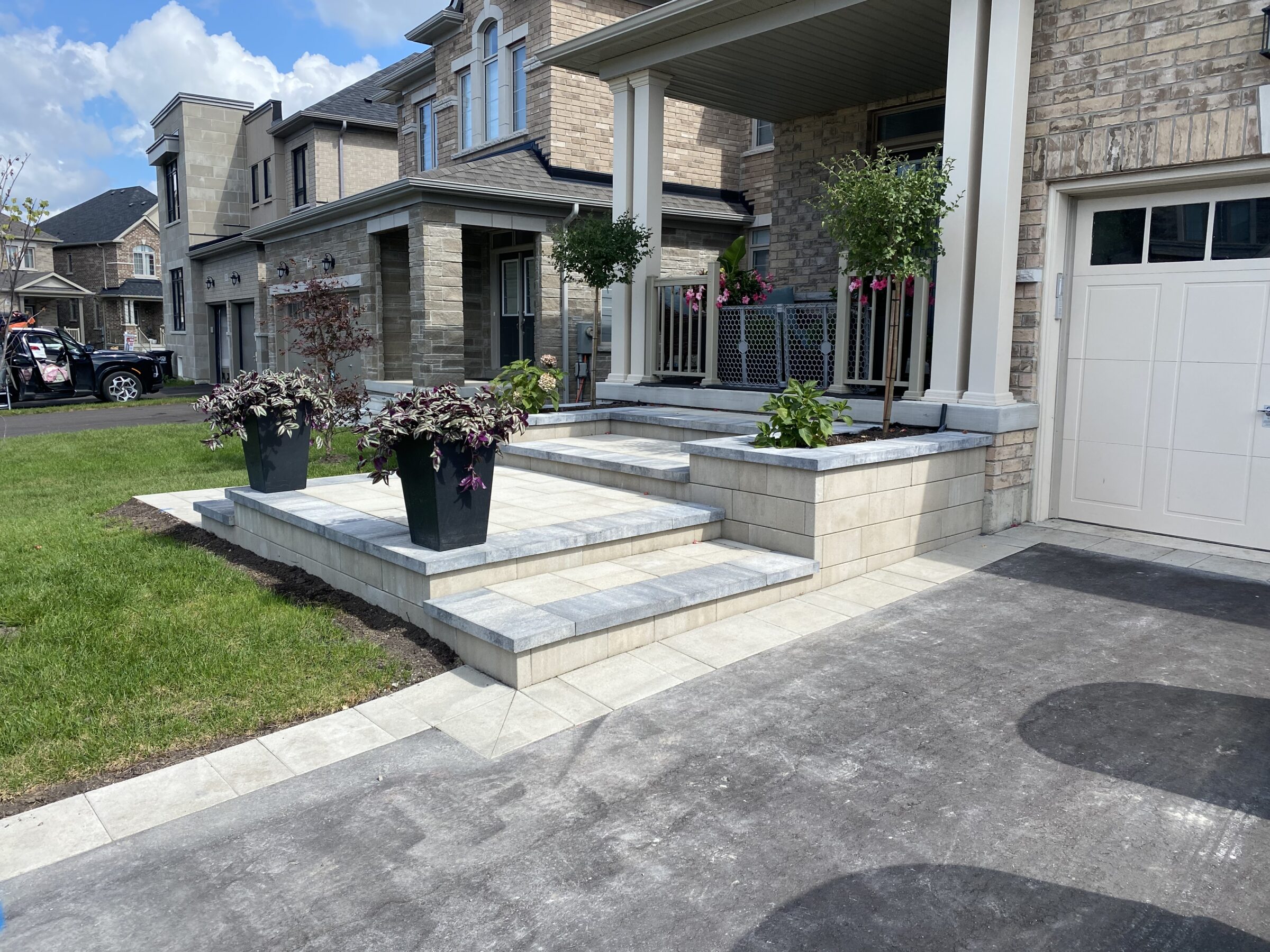 Suburban house with brick facade, elevated porch, potted plants, and a neatly paved driveway. Modern architecture with clean landscaping under a blue sky.