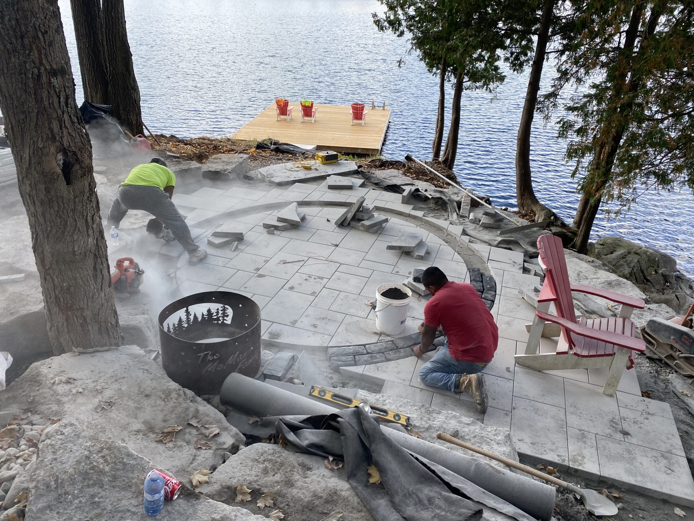 Two people work on a stone patio by a lake. Adirondack chairs and a dock are nearby, with trees surrounding the scene.