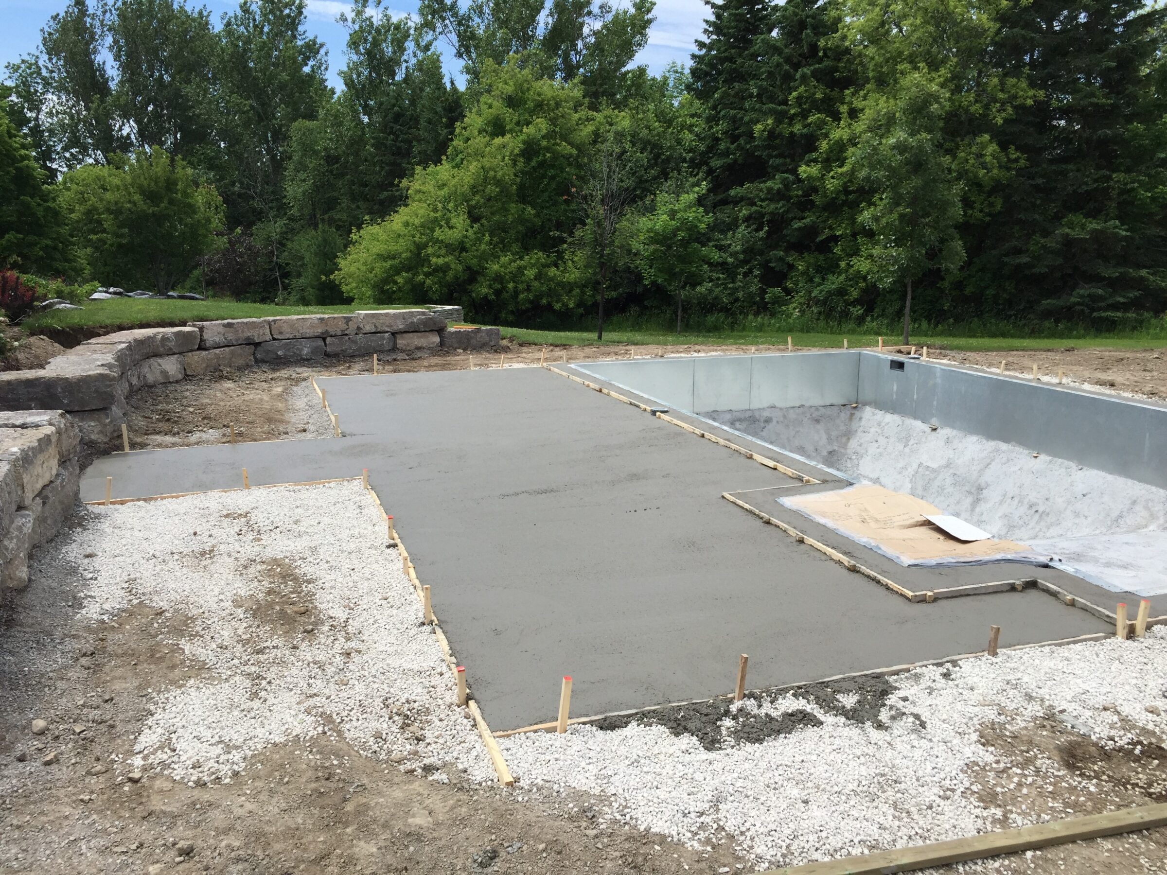 A partially constructed swimming pool in a yard, surrounded by trees and stone walls, with concrete and gravel surfaces around the edge.