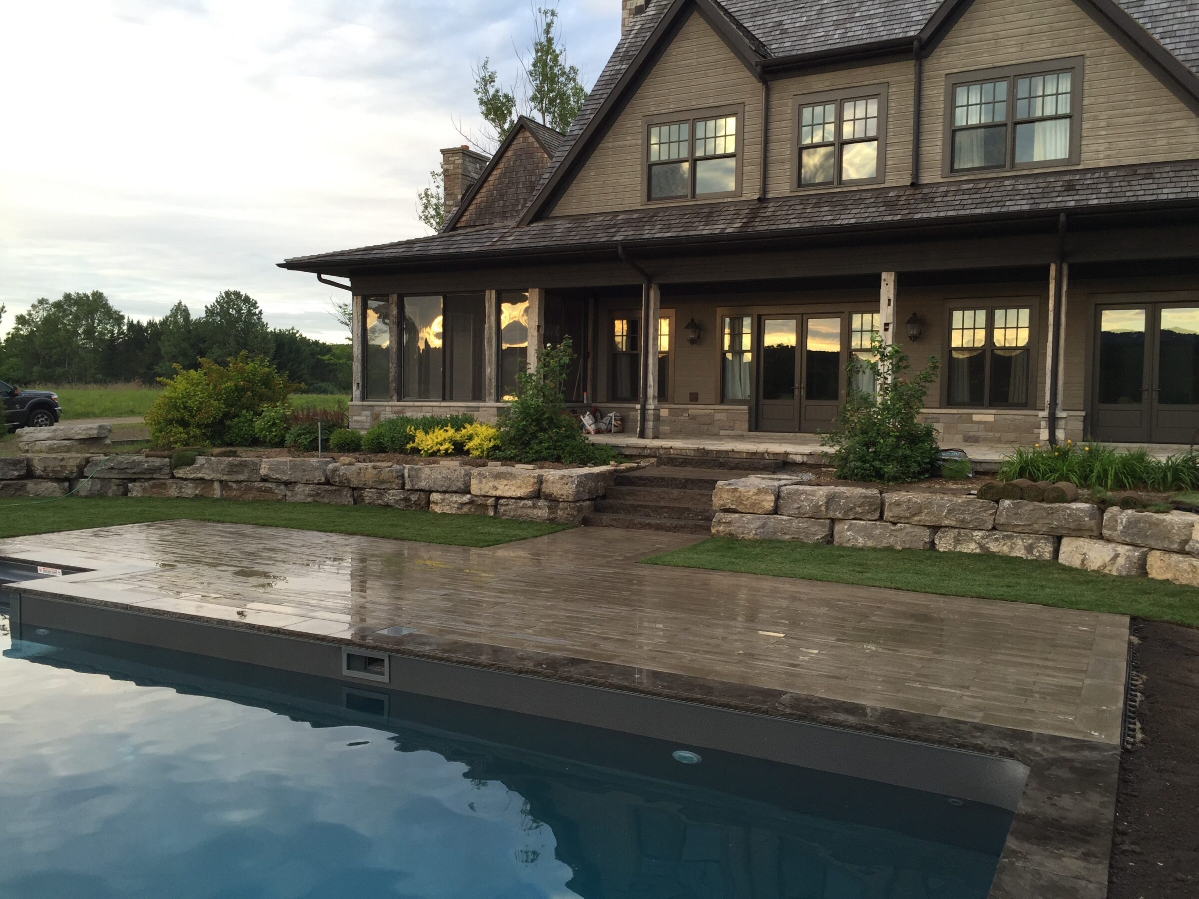 A serene countryside house with large windows, rustic stone patio, landscaped garden, and reflecting swimming pool under a partly cloudy sky.