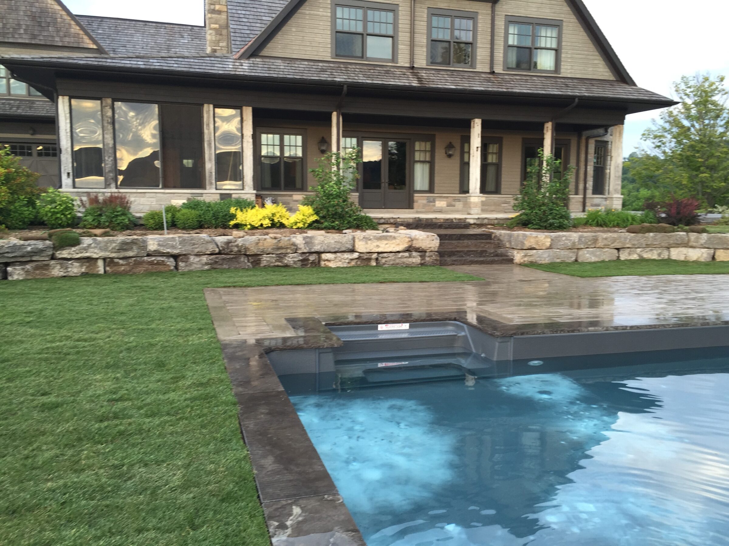 A modern house with a porch is surrounded by a green lawn. An outdoor pool is in the foreground, reflecting clouded skies.