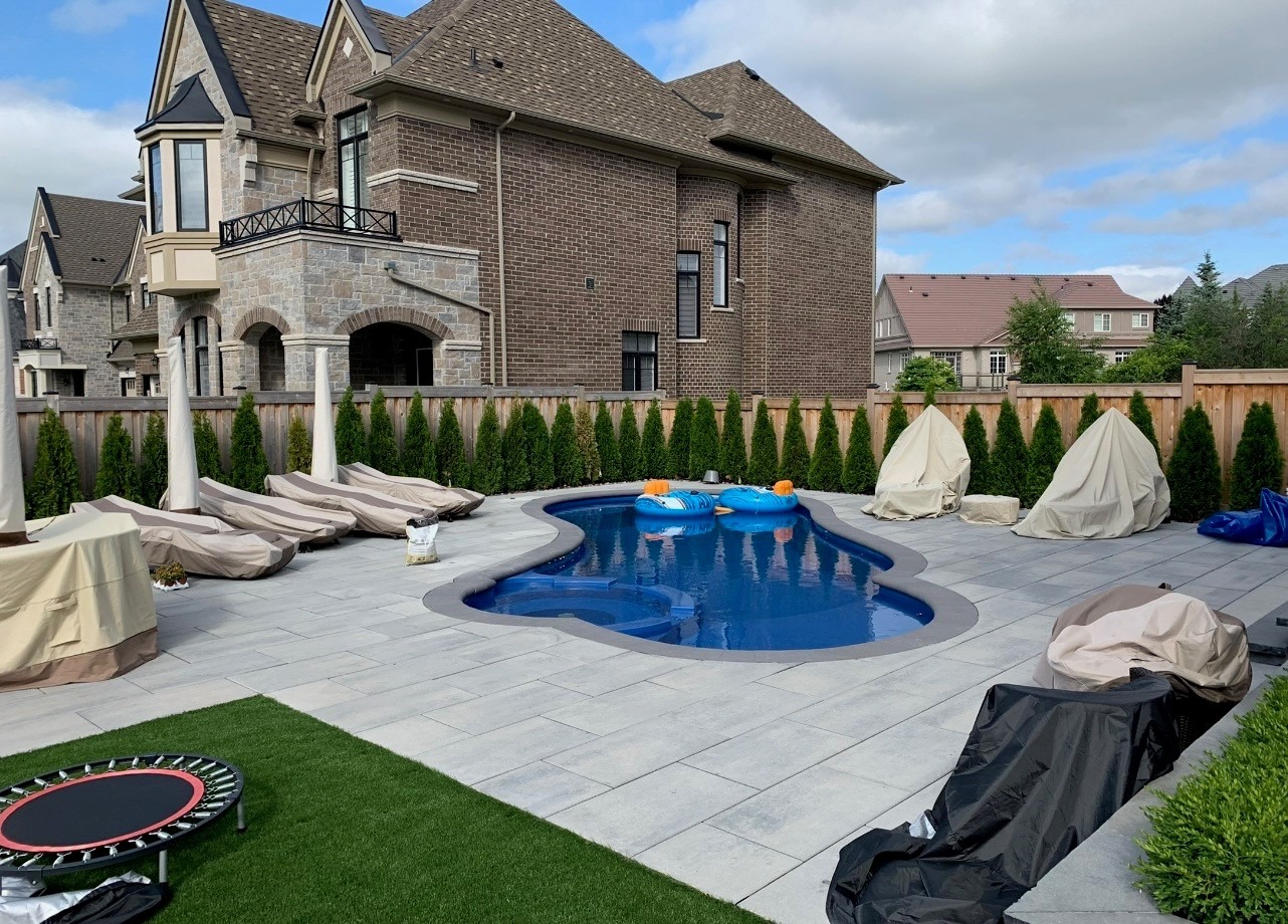 Elegant backyard features a small pool, covered patio furniture, and manicured lawn, surrounded by brick houses. The sky is partly cloudy.