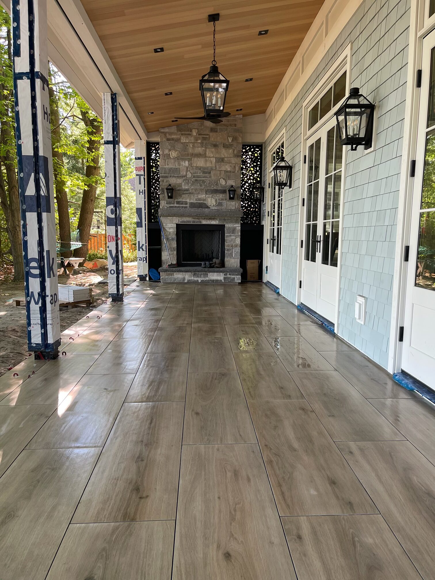 A covered outdoor patio with wood-like tiles, stone fireplace, hanging lanterns, and surrounding forest visible through pillars. No people present.