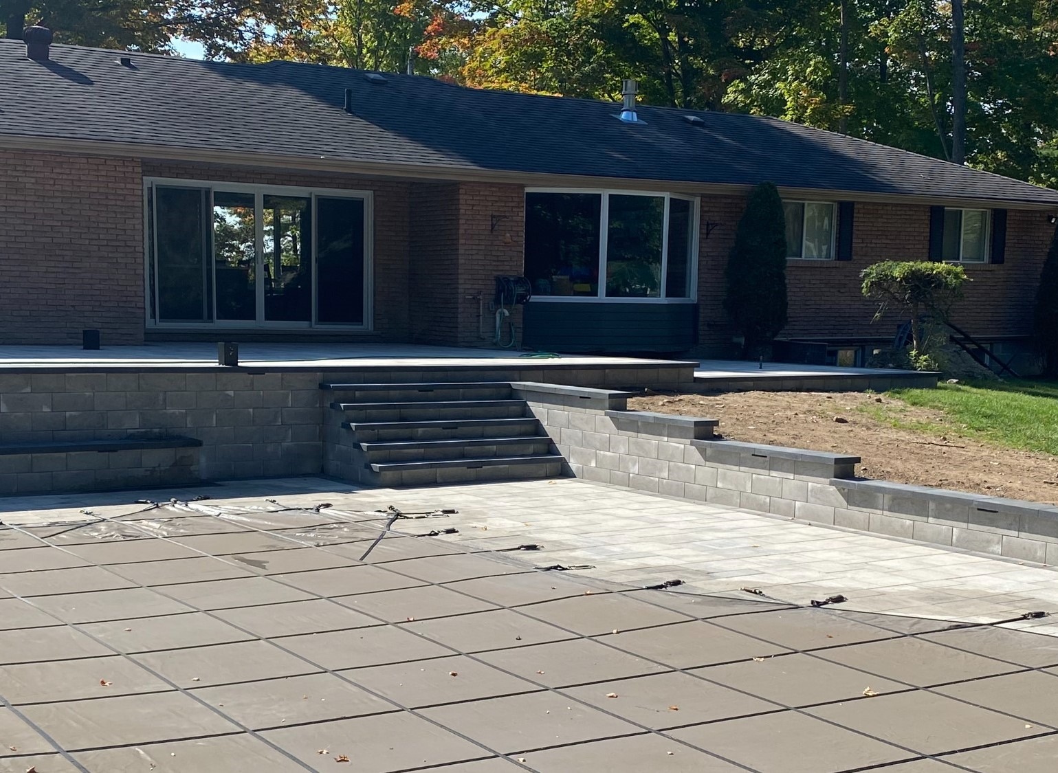 A brick house with sliding glass doors opens onto a spacious backyard patio with stairs, surrounded by trees and lawn.