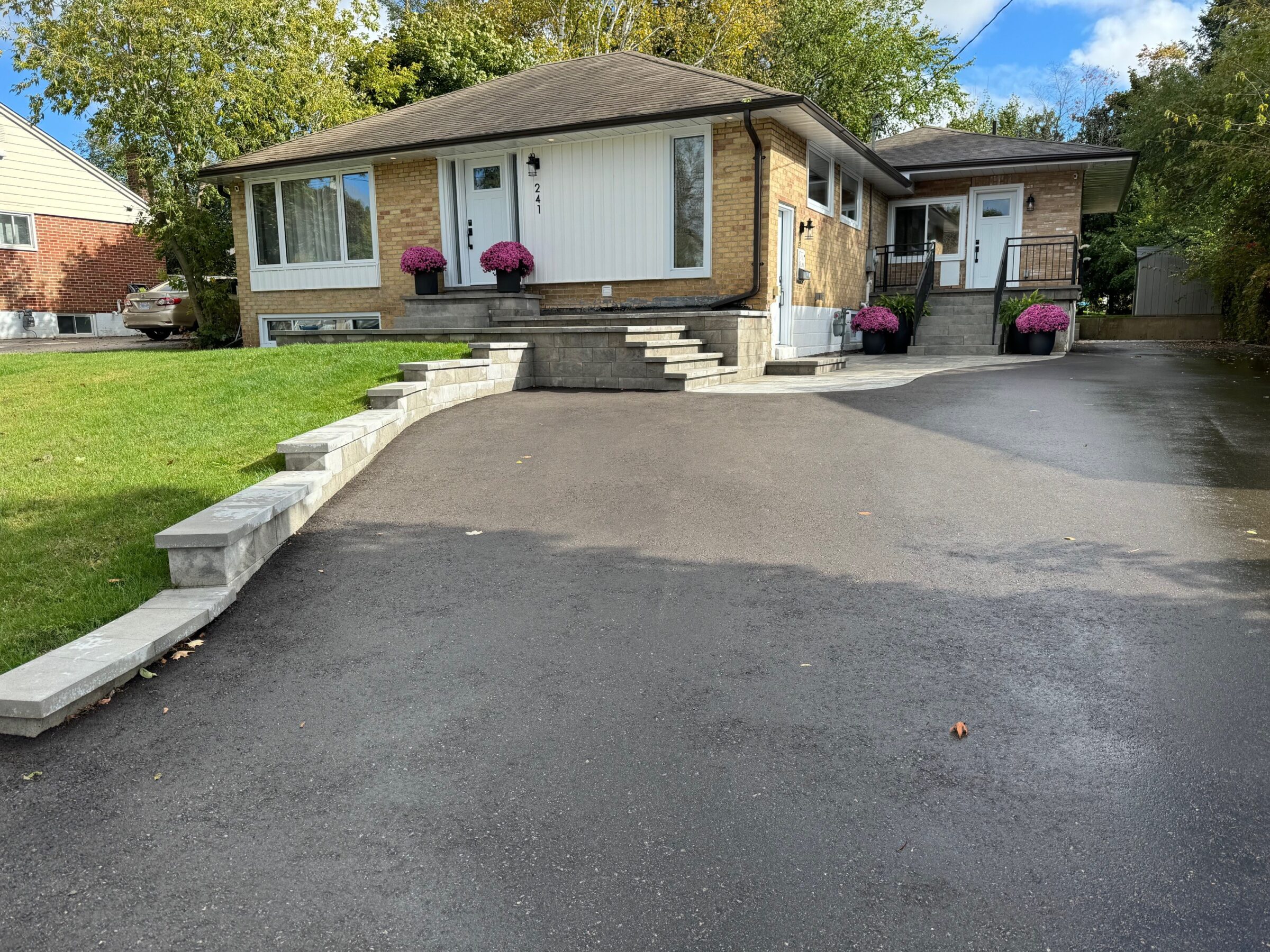 A brick house with a paved driveway and steps, surrounded by grass and trees, featuring potted plants by the entrance.