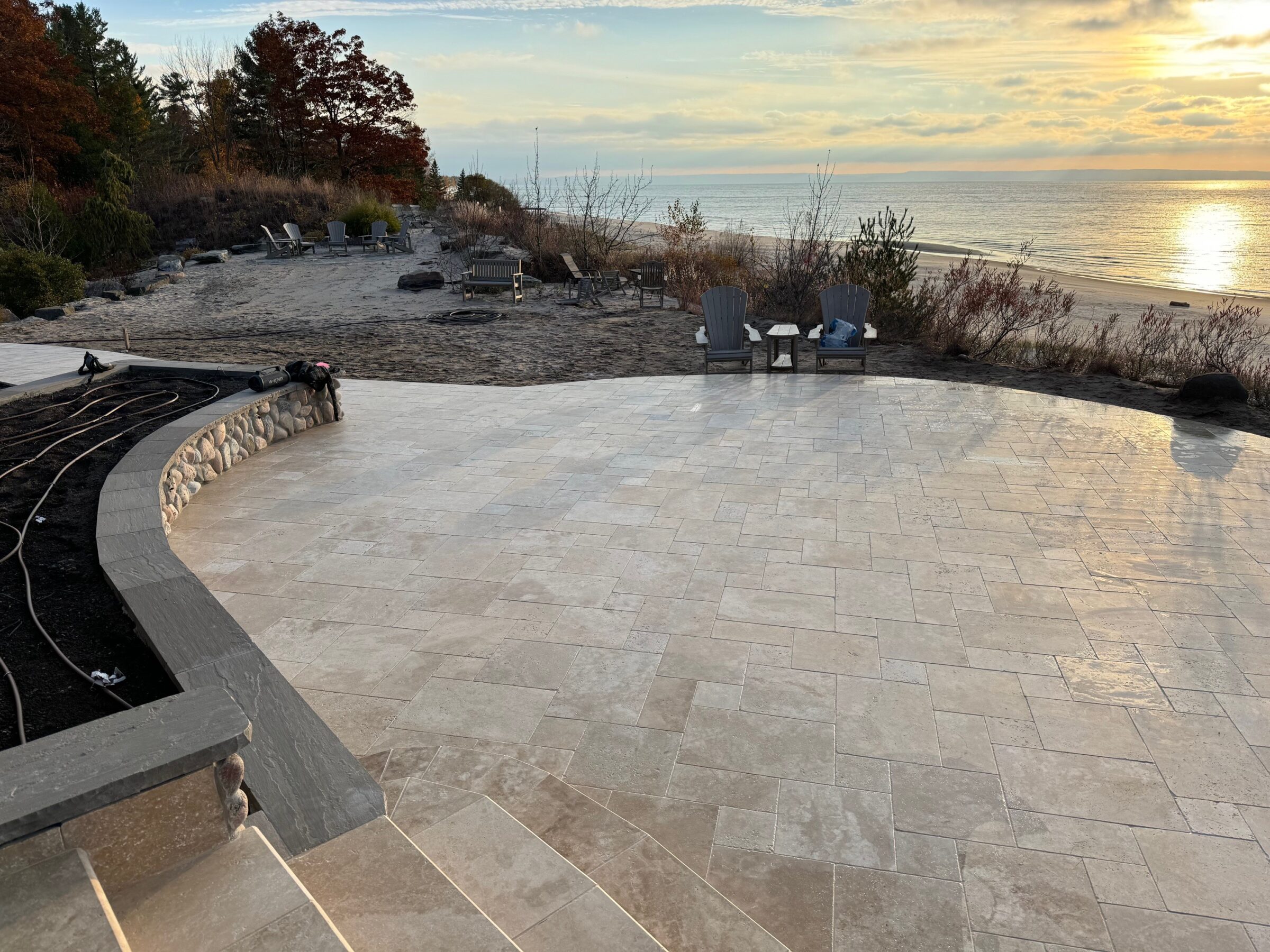 A scenic view of a beachside patio with stone tiles, overlooking the ocean during sunset. Two chairs face the water, surrounded by nature.