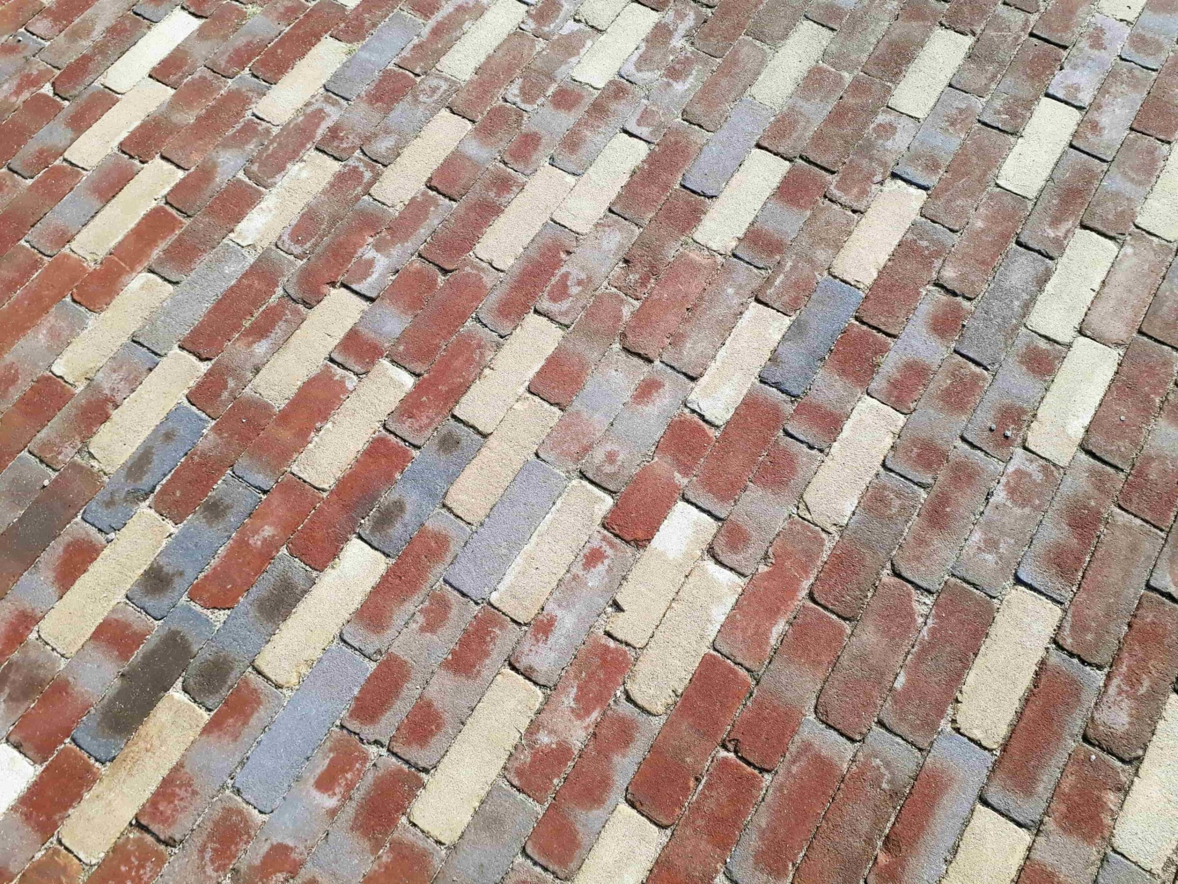 A close-up view of a paved surface with alternating reddish, beige, and grey bricks arranged in a herringbone pattern. No people or landmarks visible.