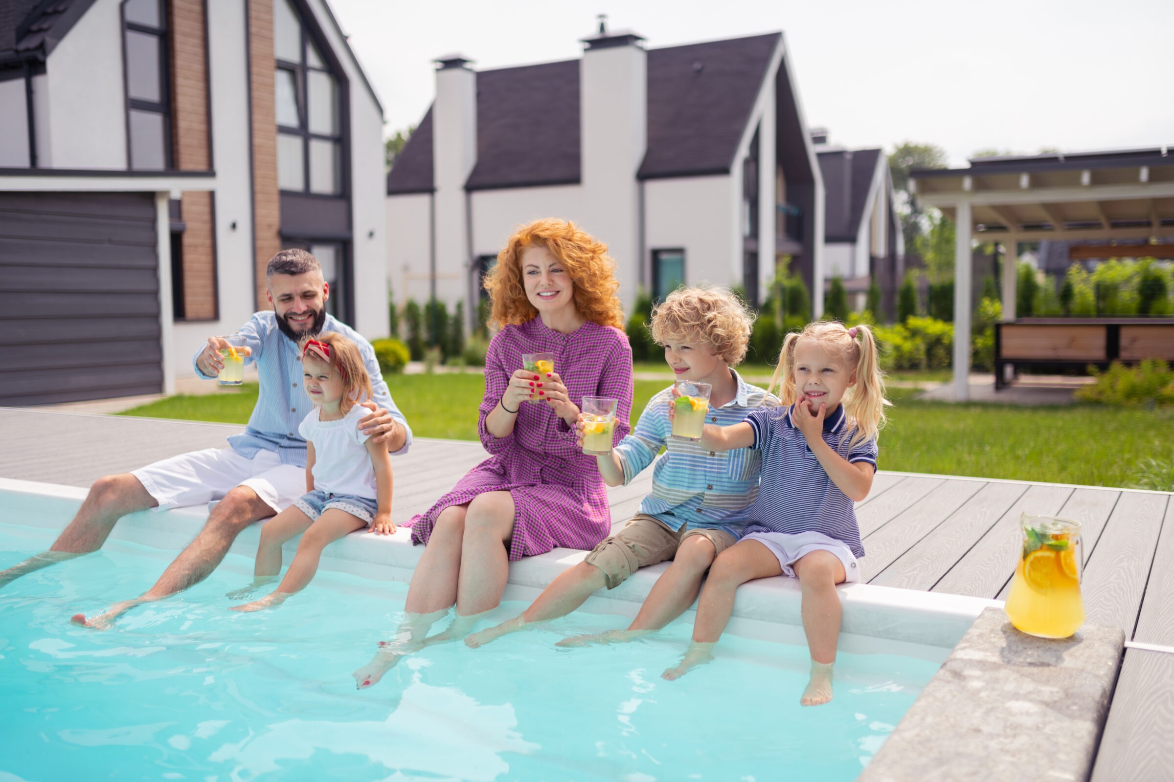 A family of five relaxes by a modern pool, enjoying drinks, with contemporary houses in the background on a sunny day.