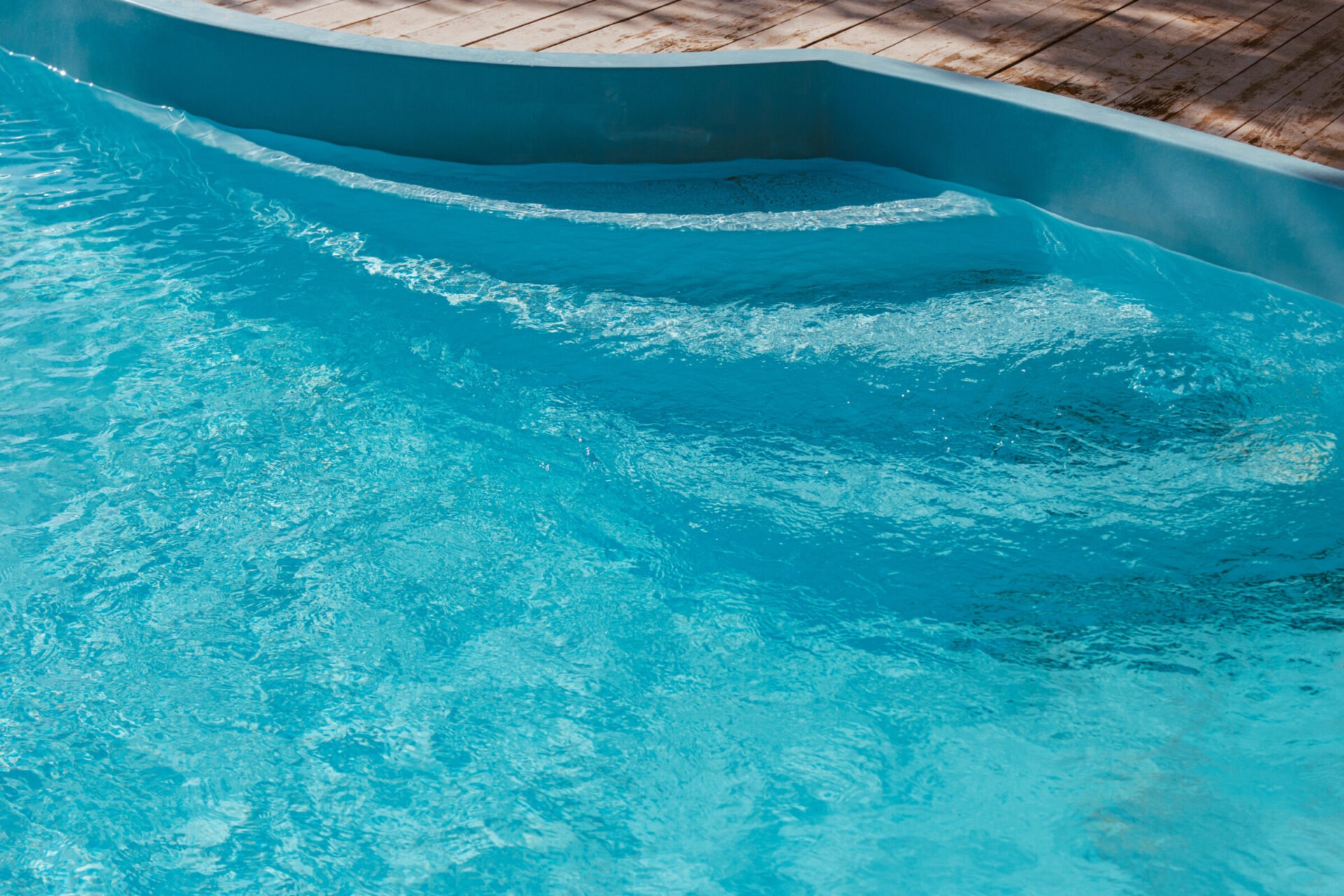 Clear blue water in a swimming pool with curved steps, bordered by a wooden deck. Sunlight gently reflects on the surface.