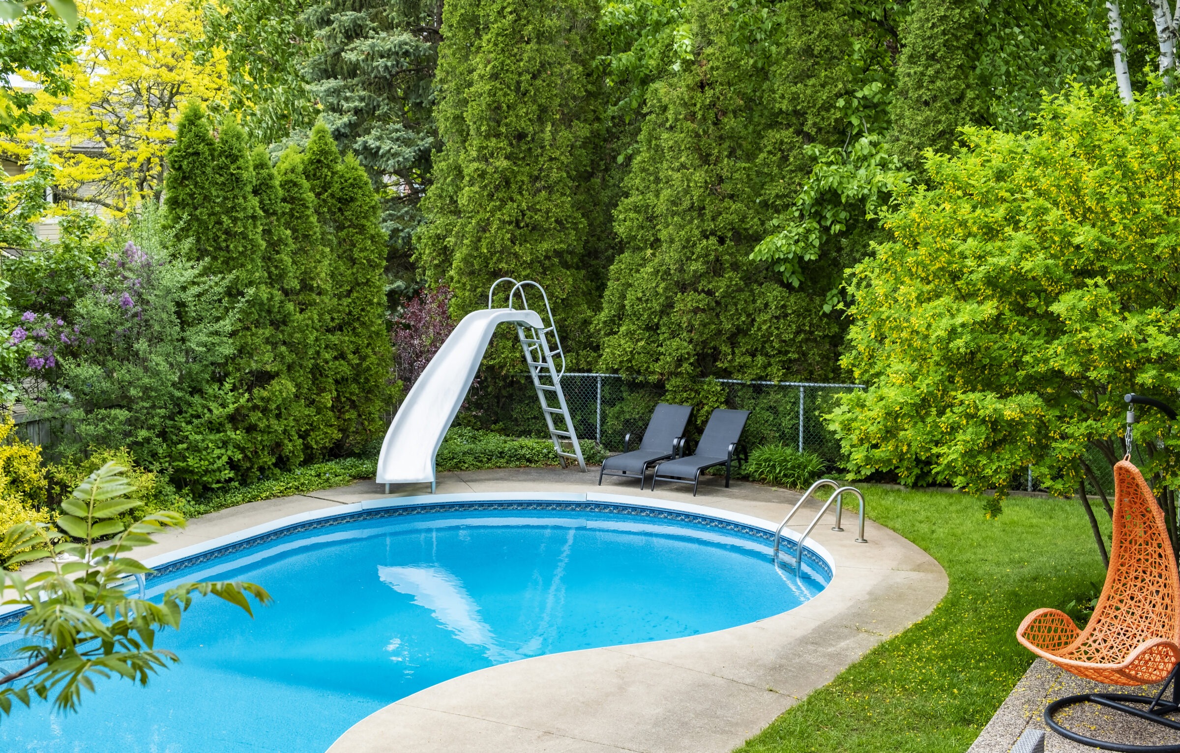 A backyard with a kidney-shaped pool, slide, and two loungers, surrounded by lush greenery and trees, creating a relaxing outdoor oasis.