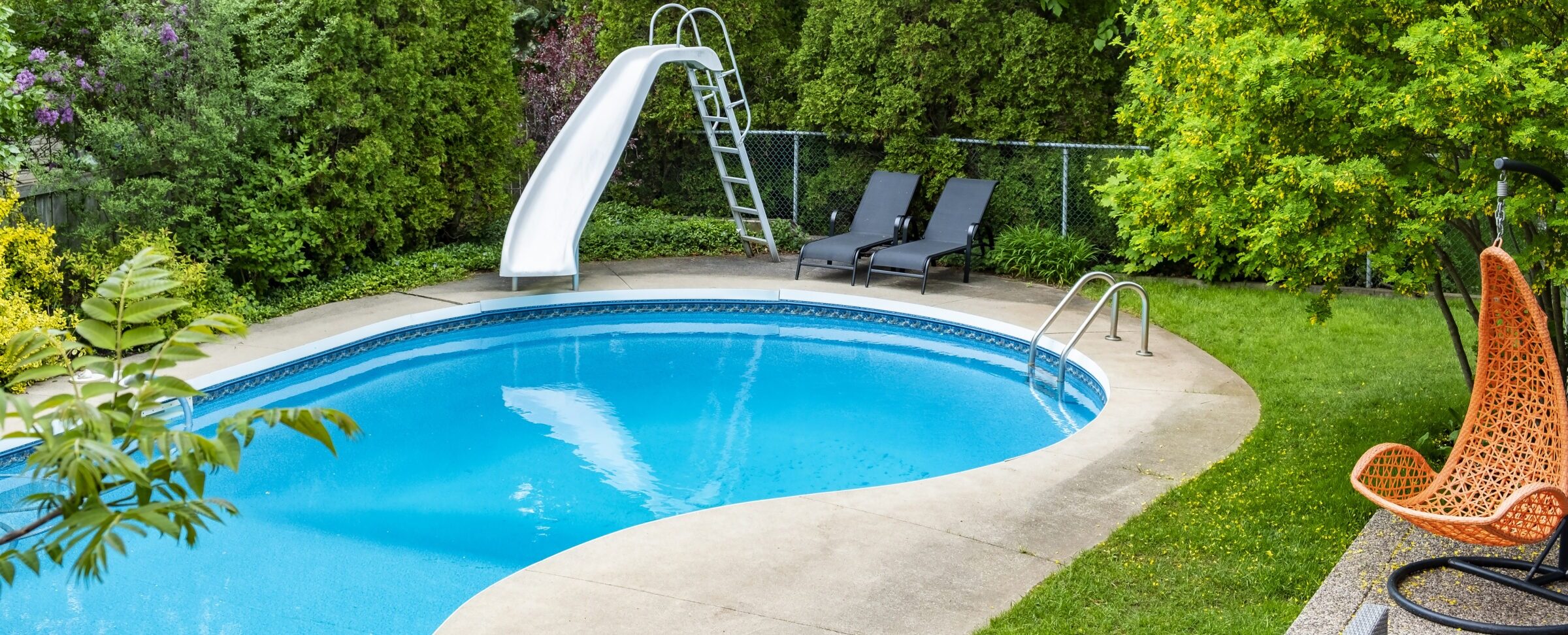 A backyard pool with a slide, surrounded by greenery and two lounge chairs on a sunny day.
