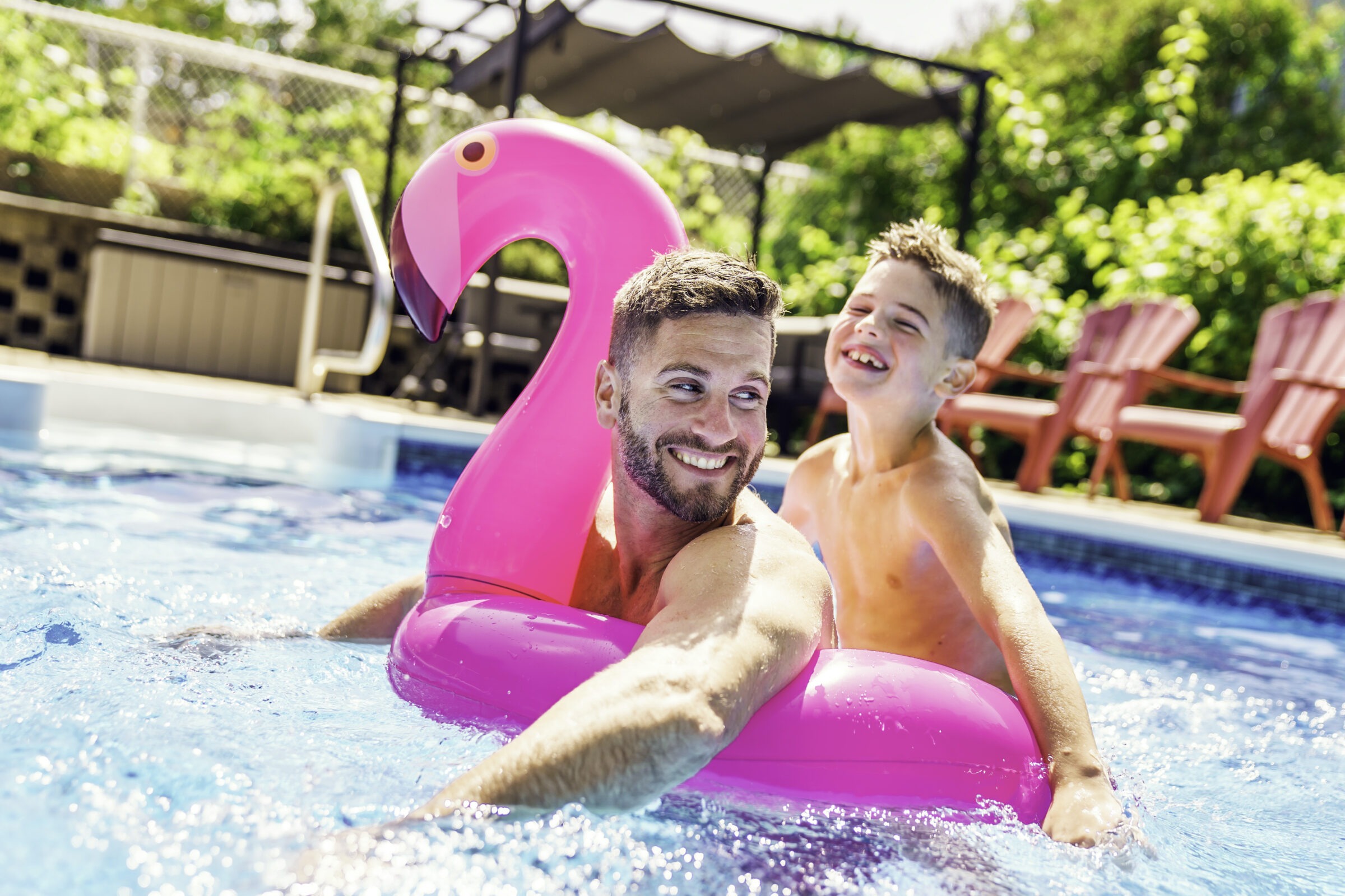 A person and child enjoy swimming with a pink inflatable flamingo in a sunny pool surrounded by greenery and outdoor furniture.