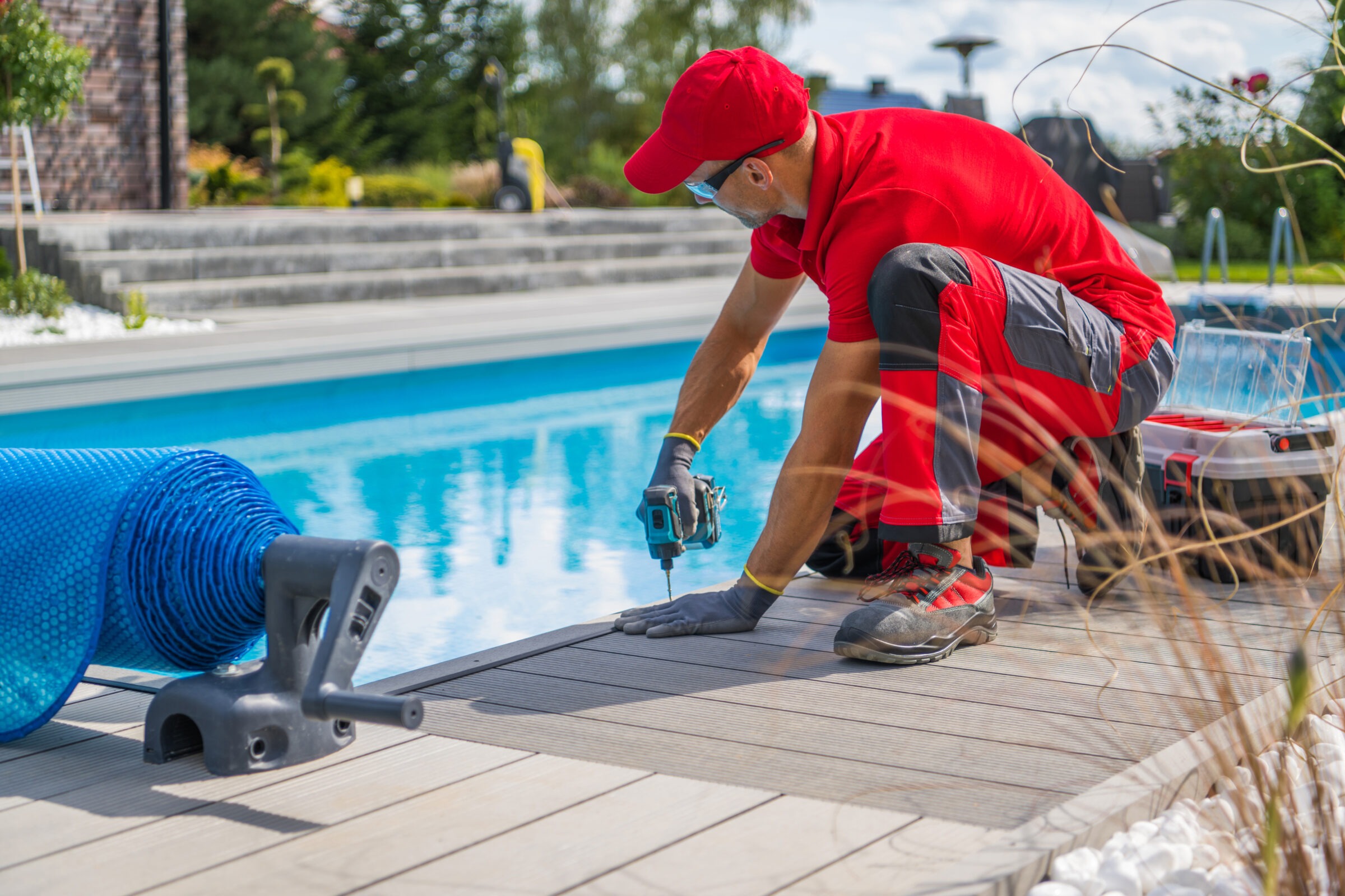 A person in red attire uses a power drill near a modern swimming pool, with a pool cover nearby, on a sunny day.