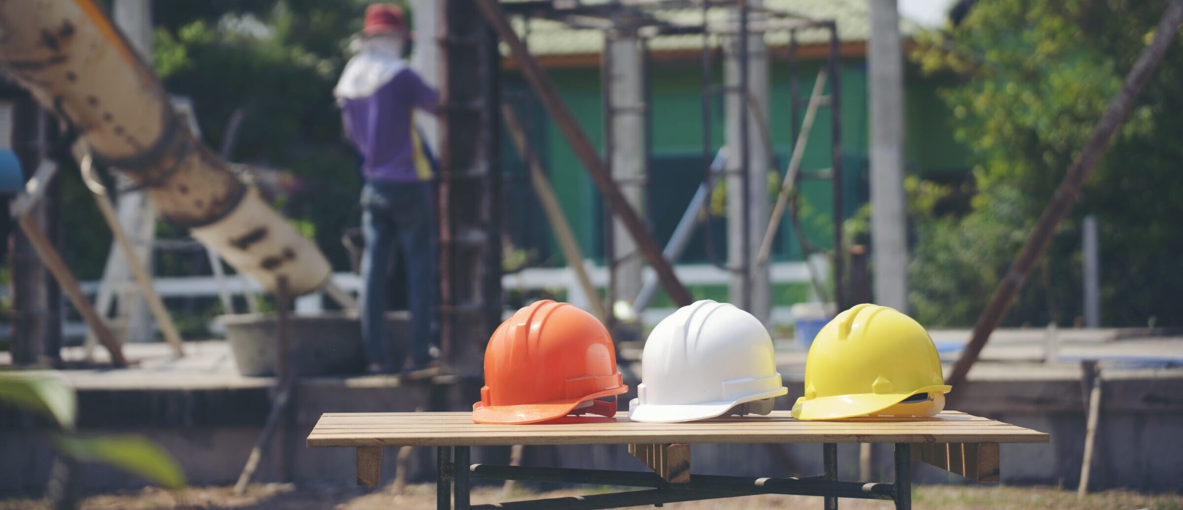 Three construction helmets on a table at a building site, with a person working in the background wearing protective gear and equipment visible.
