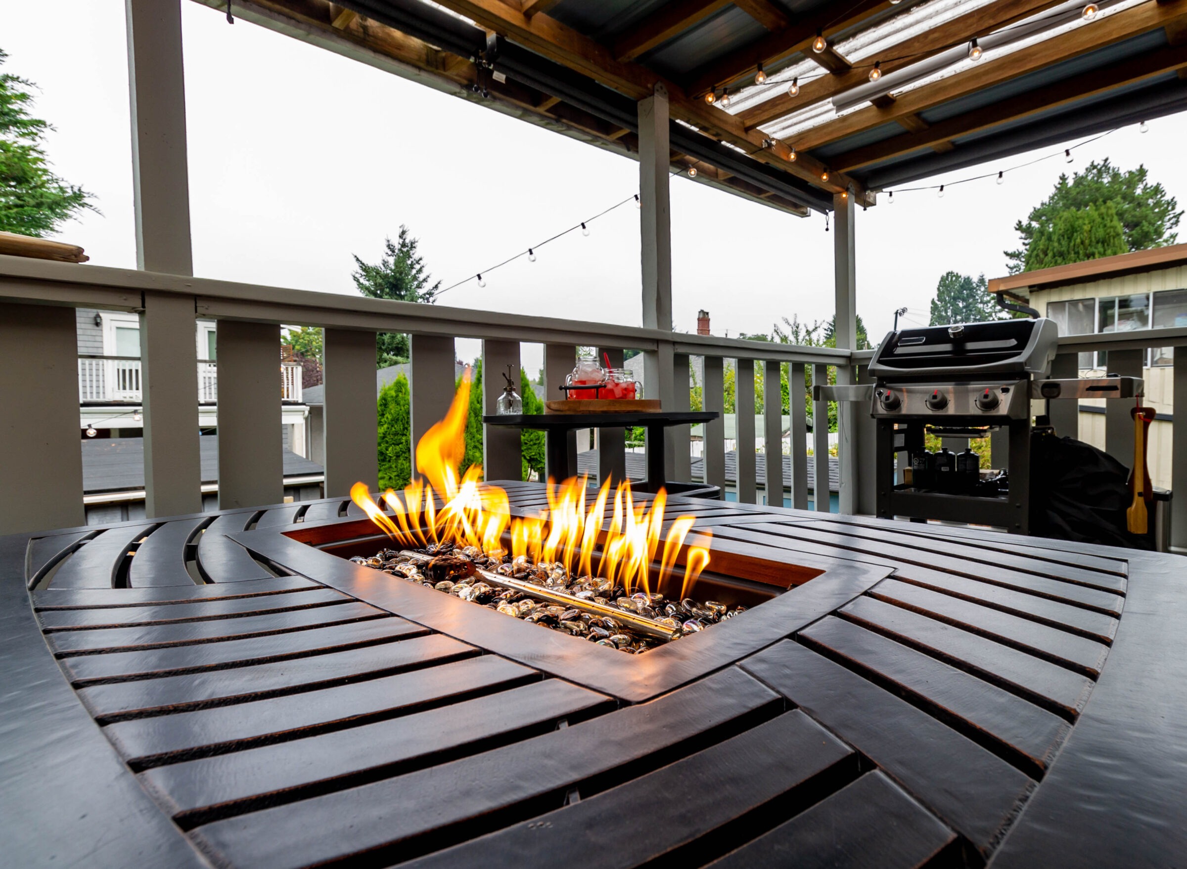 A cozy patio features a modern fire pit, string lights, and a barbecue grill, overlooking neighboring houses and lush greenery.
