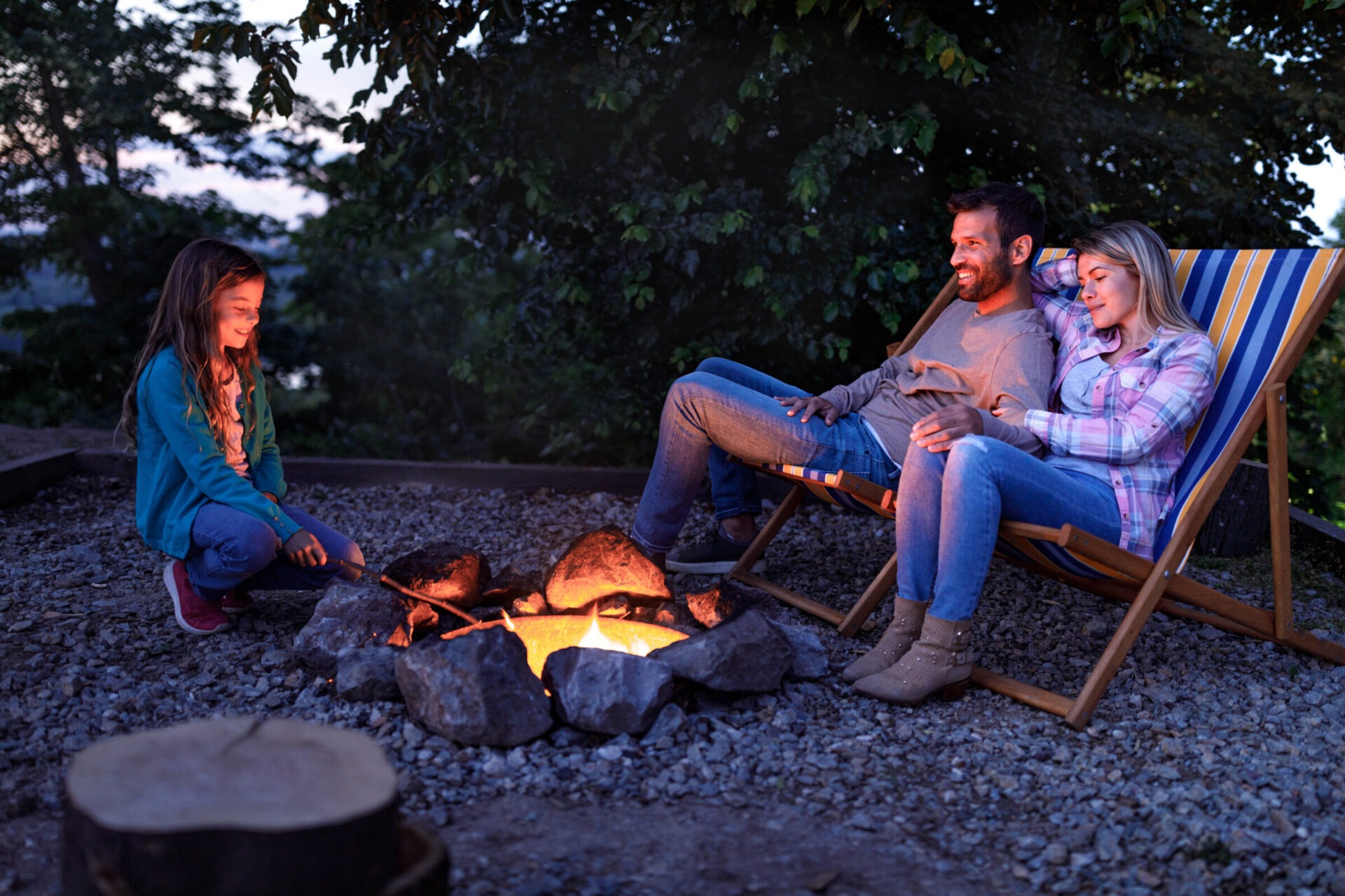Three people sitting around a campfire in a natural setting; a child toasting marshmallows, two adults relaxing in deck chairs.