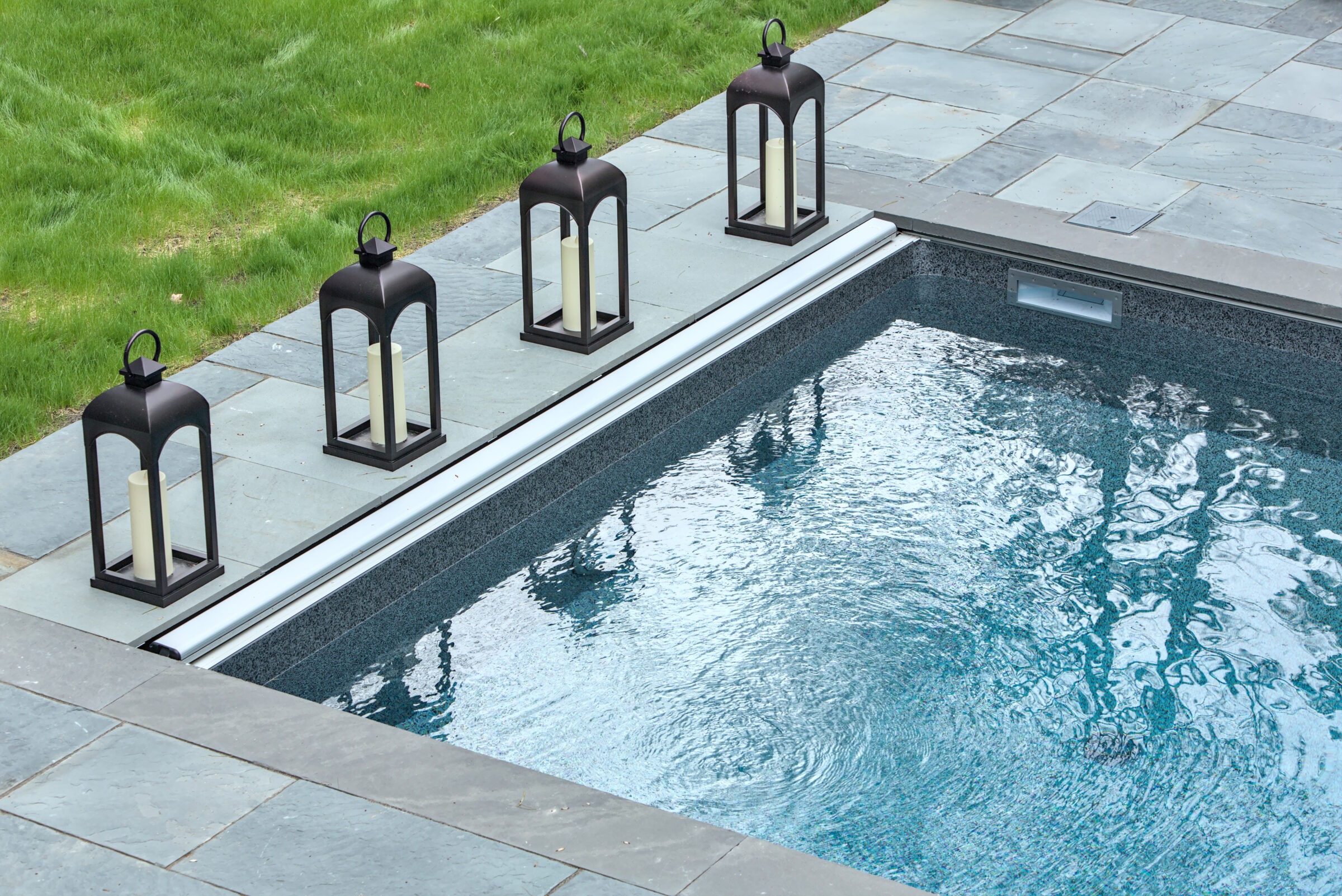 A serene poolside scene featuring four decorative lanterns, bordered by gray stone tiles and adjacent green grass.
