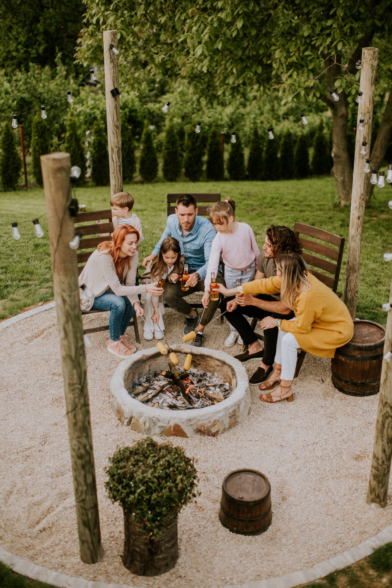 A group of seven people gather around a stone fire pit, roasting marshmallows on sticks, surrounded by greenery and string lights.