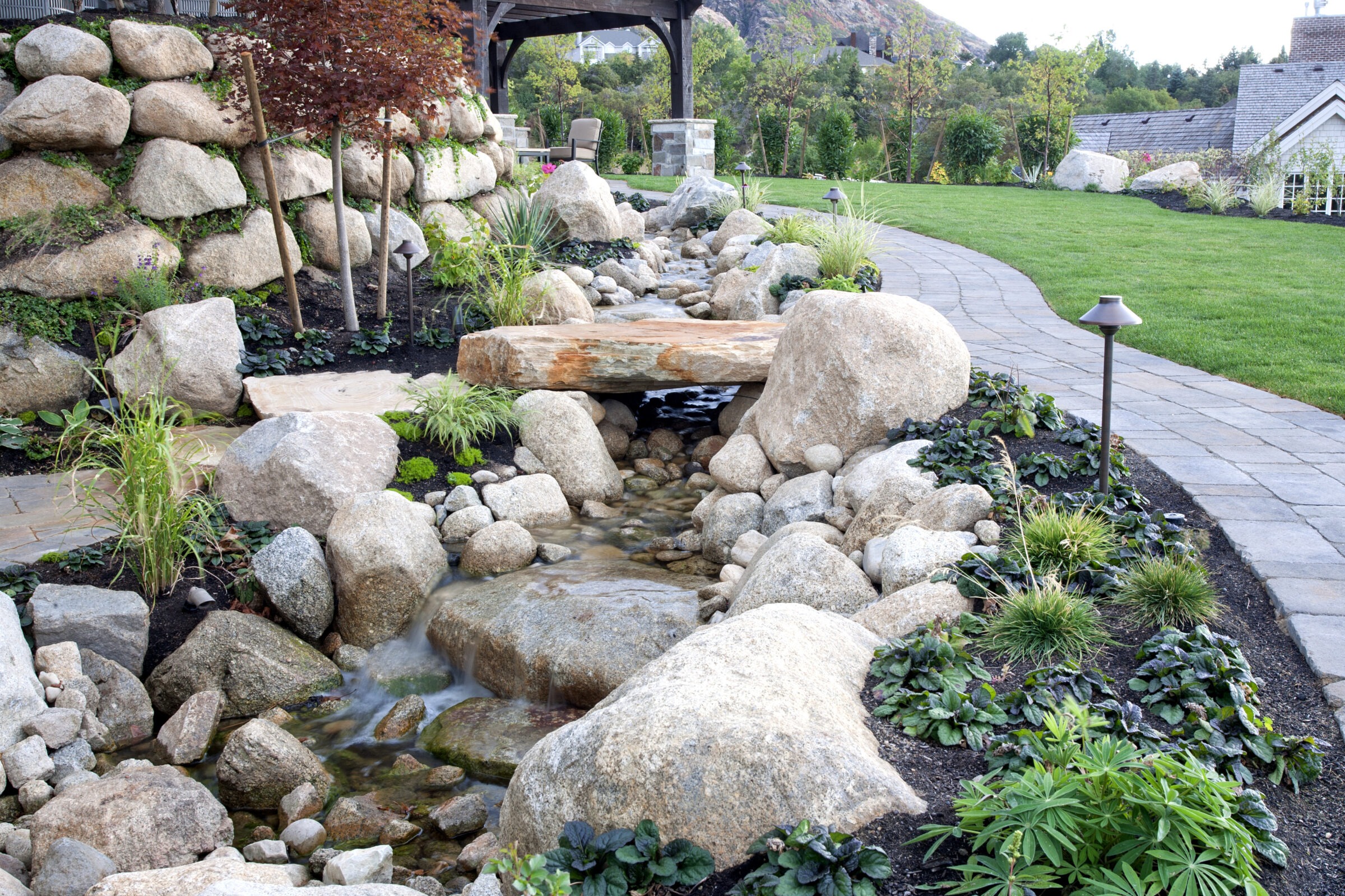 A serene garden with a stone pathway, small stream, and lush greenery. A quaint building is visible in the background.