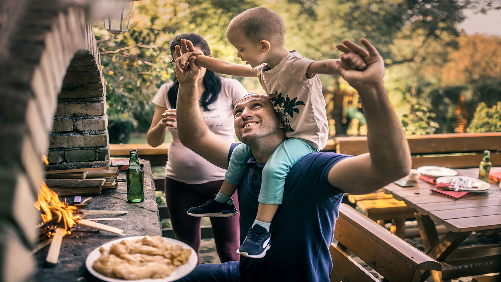 A person holds a child on shoulders near a brick grill, while another person stands nearby. A table with drinks is in the background.