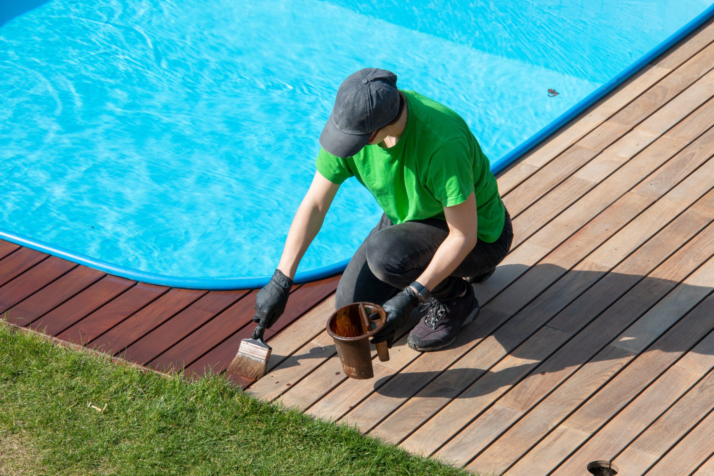 A person in a green shirt and cap paints wooden decking beside a swimming pool, with a can of paint beside them.