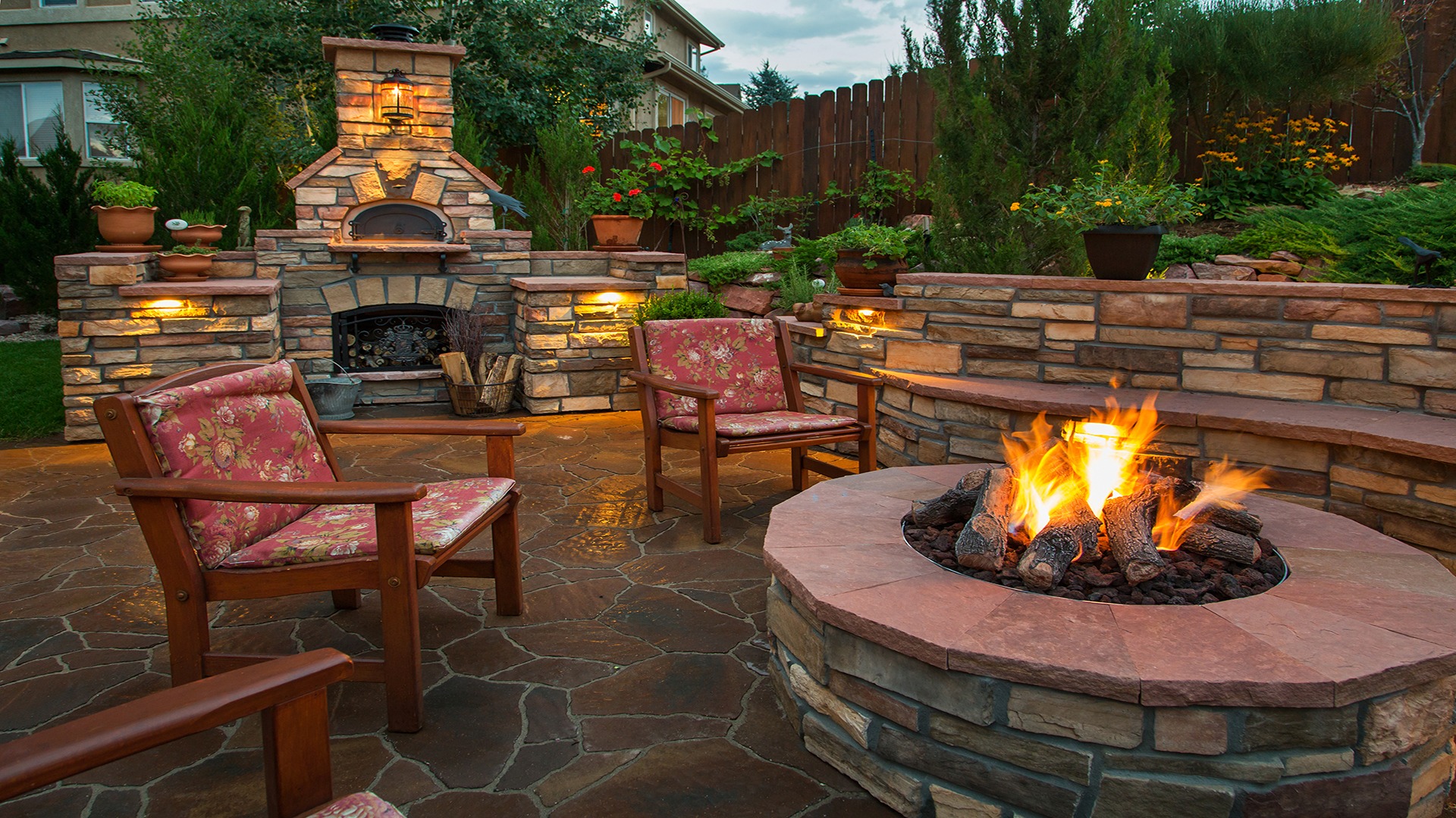Cozy backyard features a stone fire pit, wooden chairs, and a rustic outdoor oven, surrounded by lush greenery and ambient lighting.