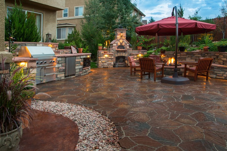 A cozy outdoor patio features a stone fireplace, grill, and seating area. Red umbrella shades chairs, surrounded by landscaped greenery and decorative lighting.