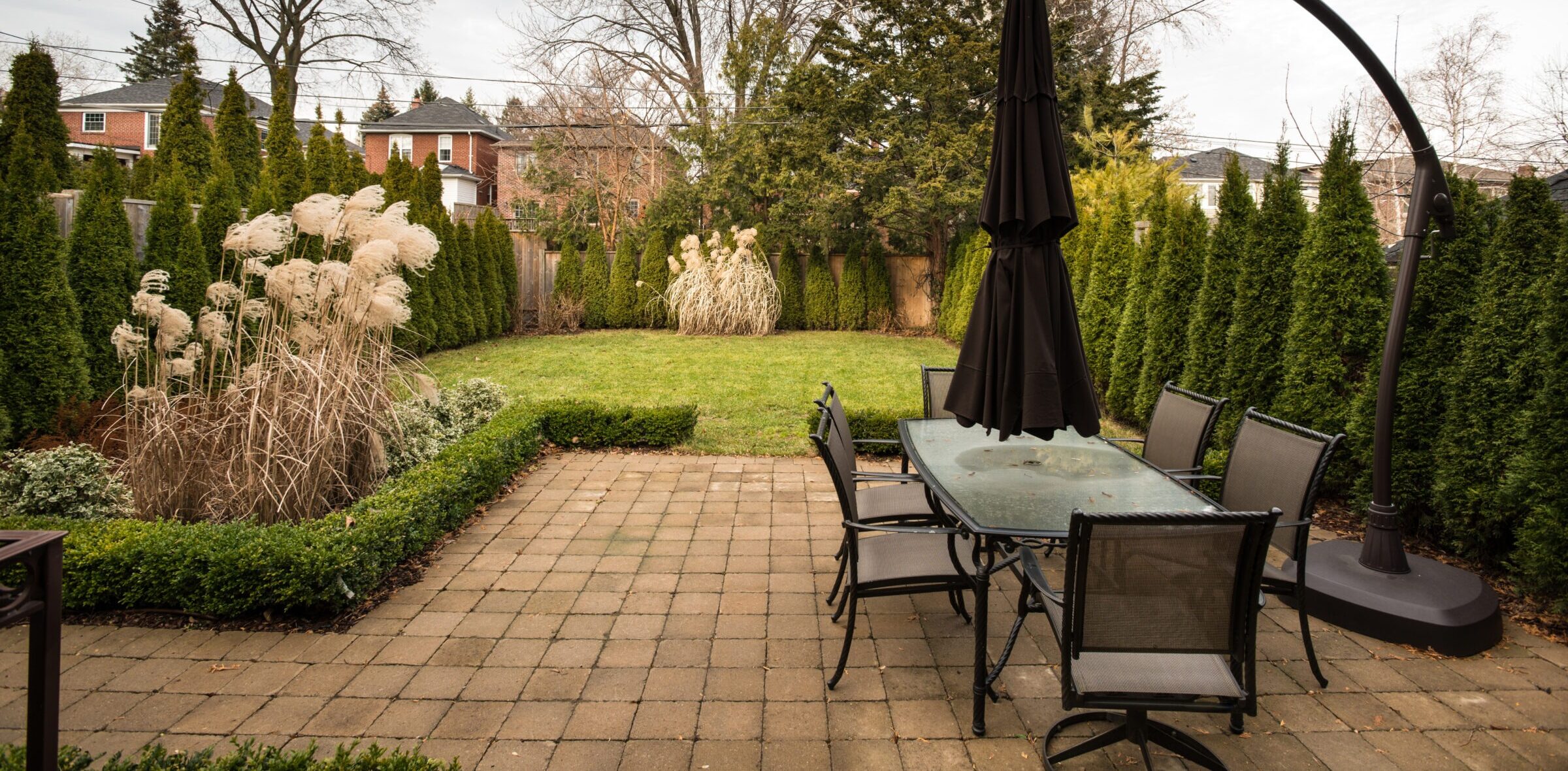 A patio with a glass table and chairs overlooks a neatly landscaped garden with tall trees and neighboring brick houses in the background.
