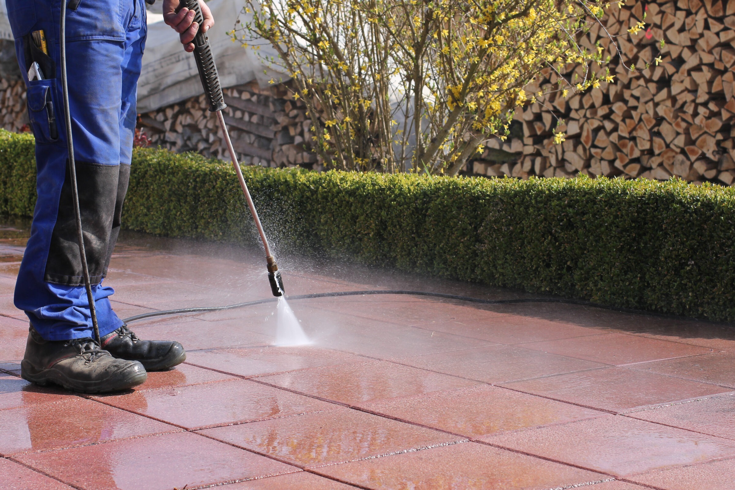 A person is pressure washing a red-tiled patio. Neatly stacked firewood and green hedges are visible in the background.