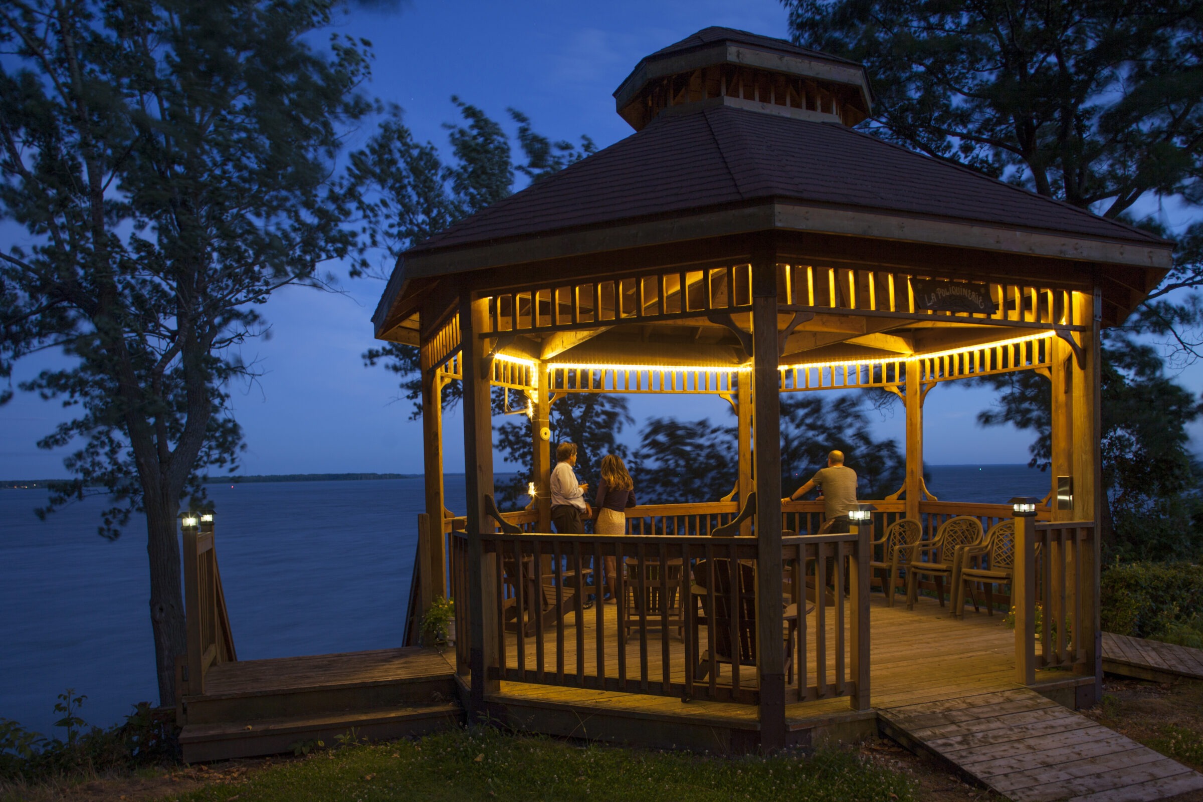 Three people stand in a lit wooden gazebo by a lake during twilight, surrounded by trees and a peaceful natural setting.