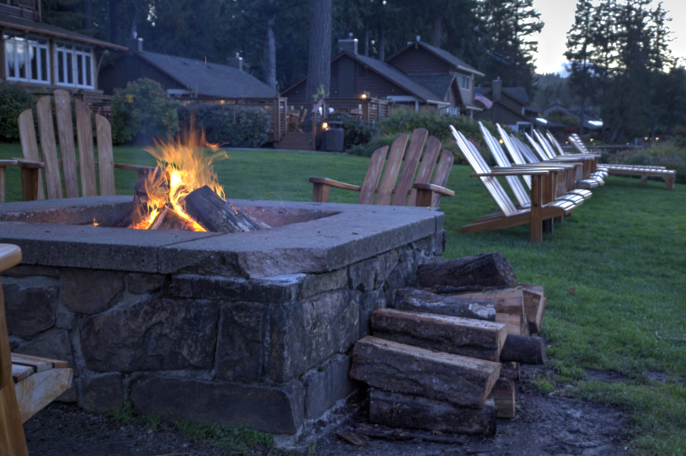 A cozy firepit with Adirondack chairs overlooks a serene grassy area, surrounded by rustic wooden cabins and tall trees at dusk.
