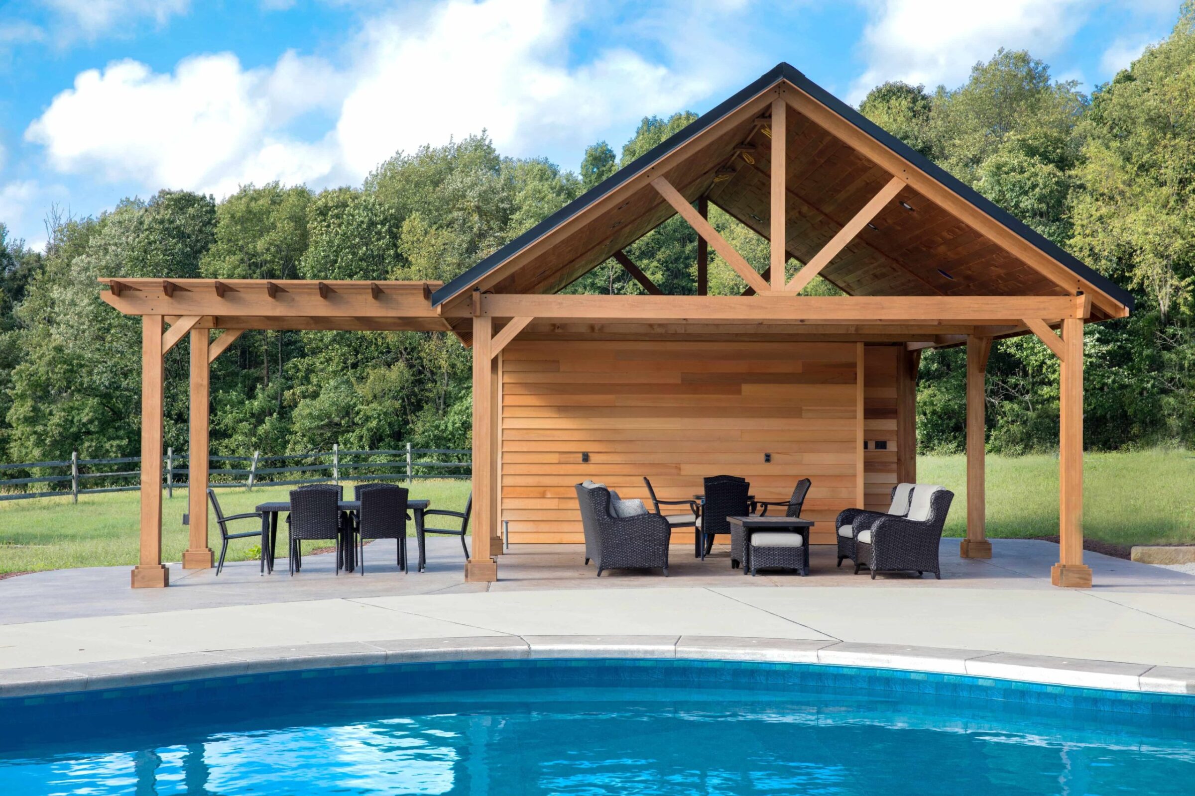 Wooden pavilion next to a clear, blue swimming pool, surrounded by trees and open grass. Outdoor furniture is arranged under the pavilion's roof.